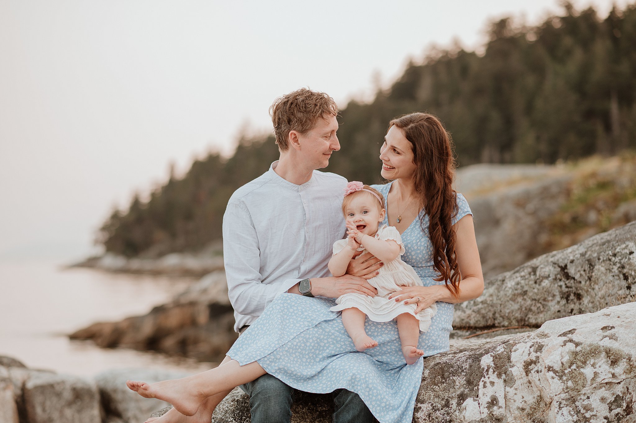 Happy parents smile at each other while sitting on a rock with their toddler daughter in mom's lap