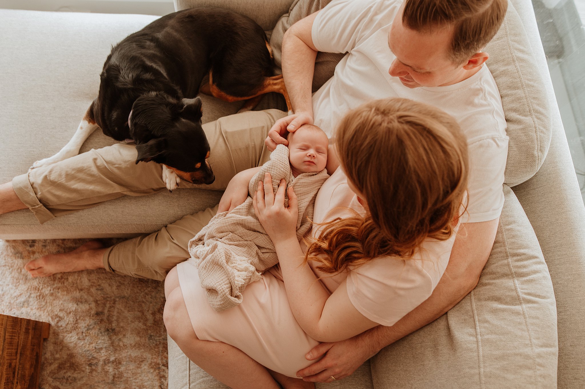 A mom and dad sit together on a couch with their dog while cradling their newborn baby in their lap