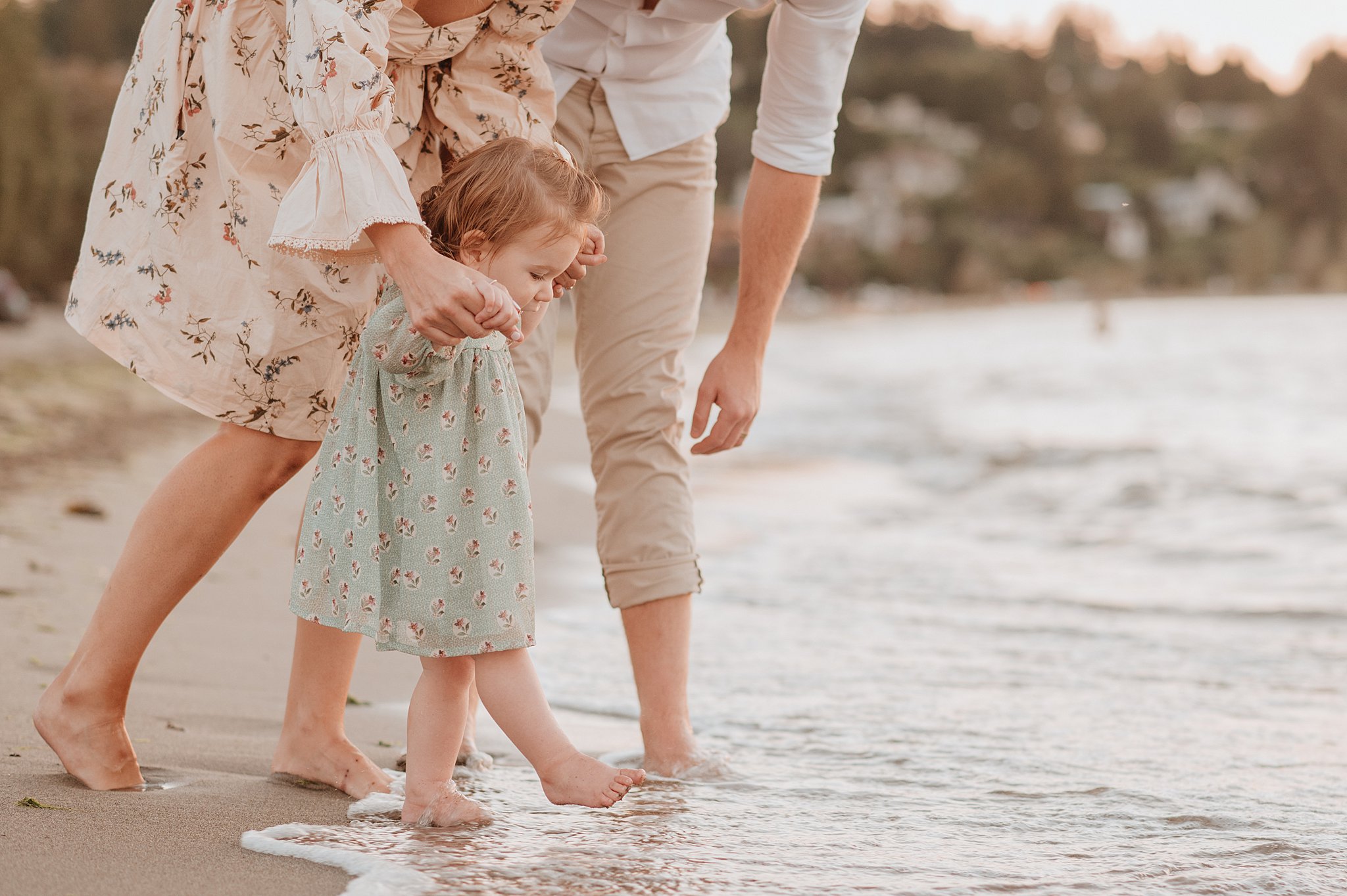 A young toddler girl in a green dress walks in the water on a beach with help from mom and dad at sunset thanks to pediatric chiropractor vancouver