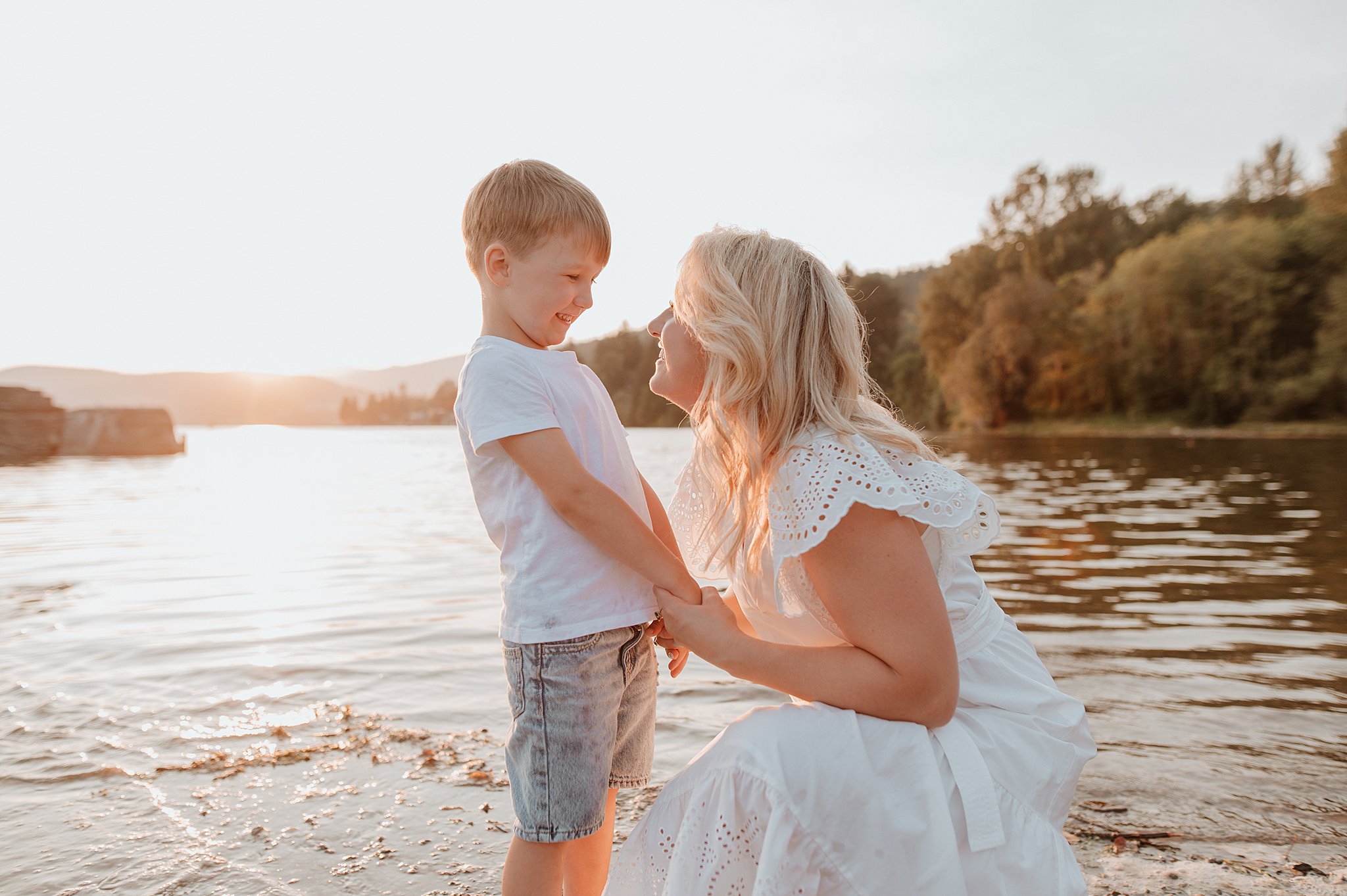 A mother in a white dress kneels down on a lake's edge to play with her toddler son at sunset
