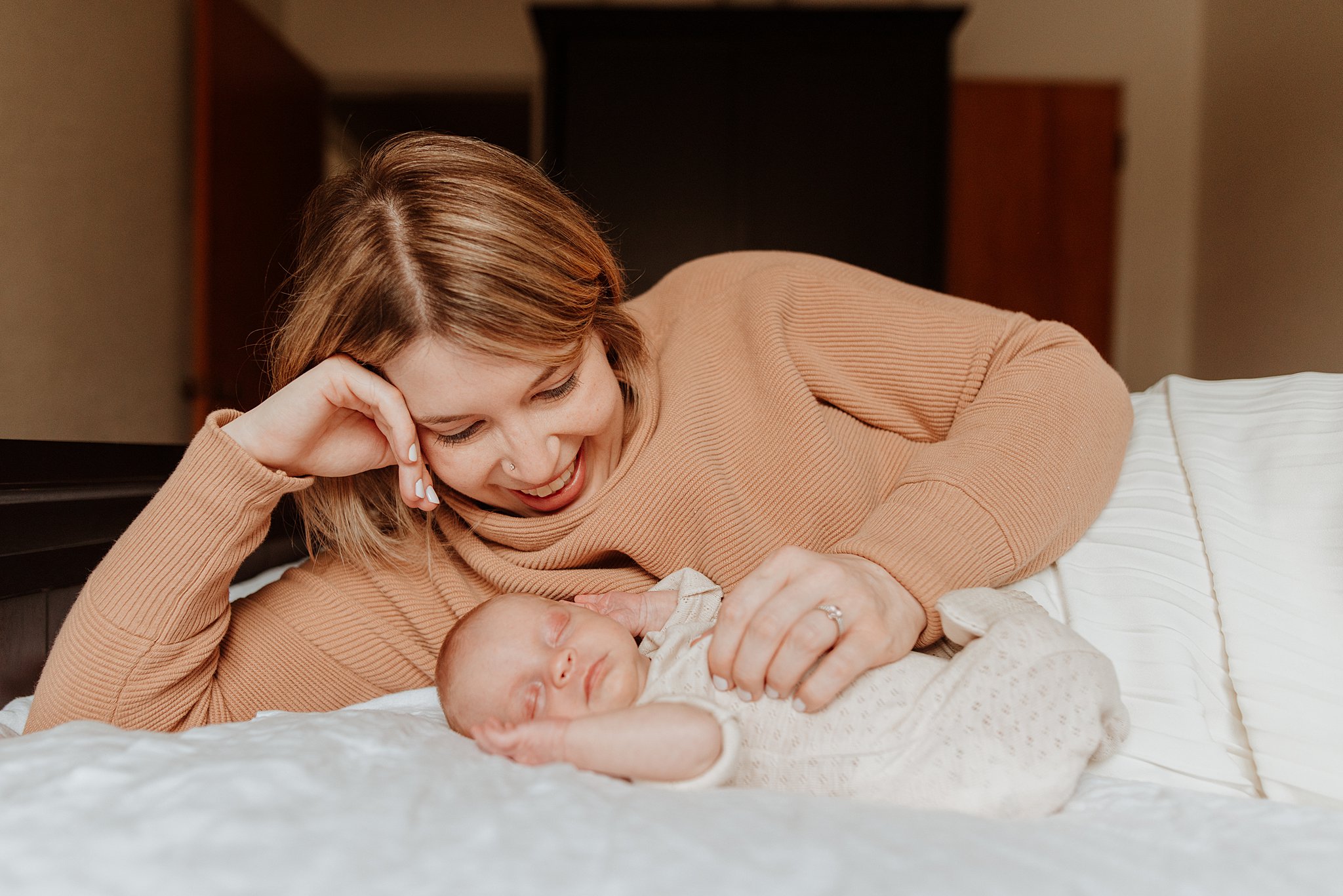 A mother in a peach sweater smiles while cuddling with her sleeping newborn baby on a bed after meeting vancouver lactation consultants