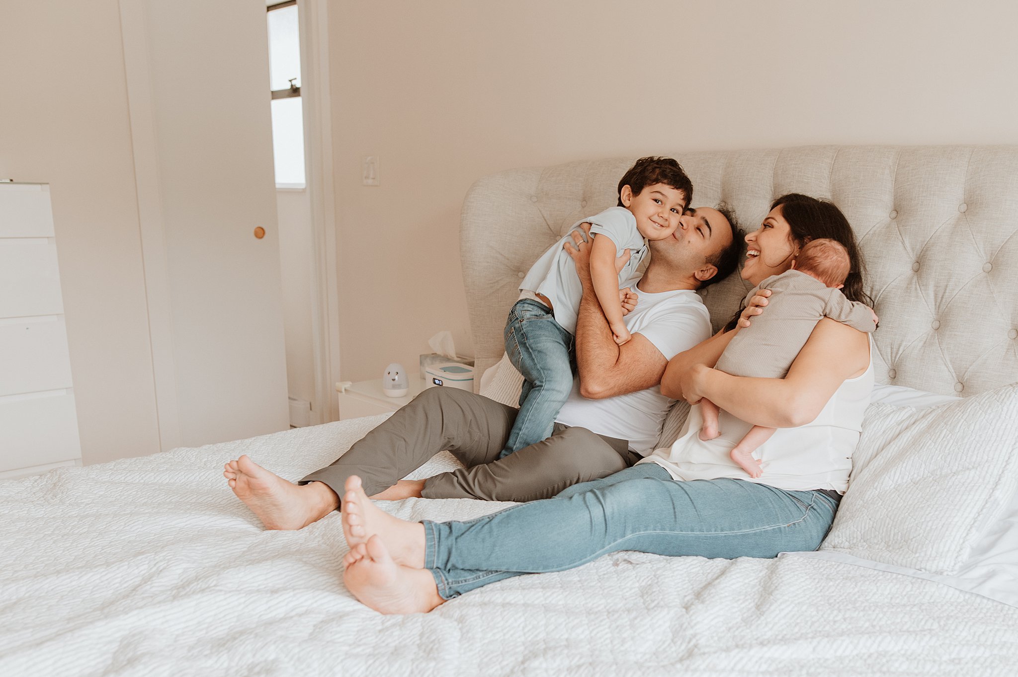 A happy mom and dad sit on their bed kissing and playing with their toddler son and newborn baby thanks to vancouver prenatal yoga