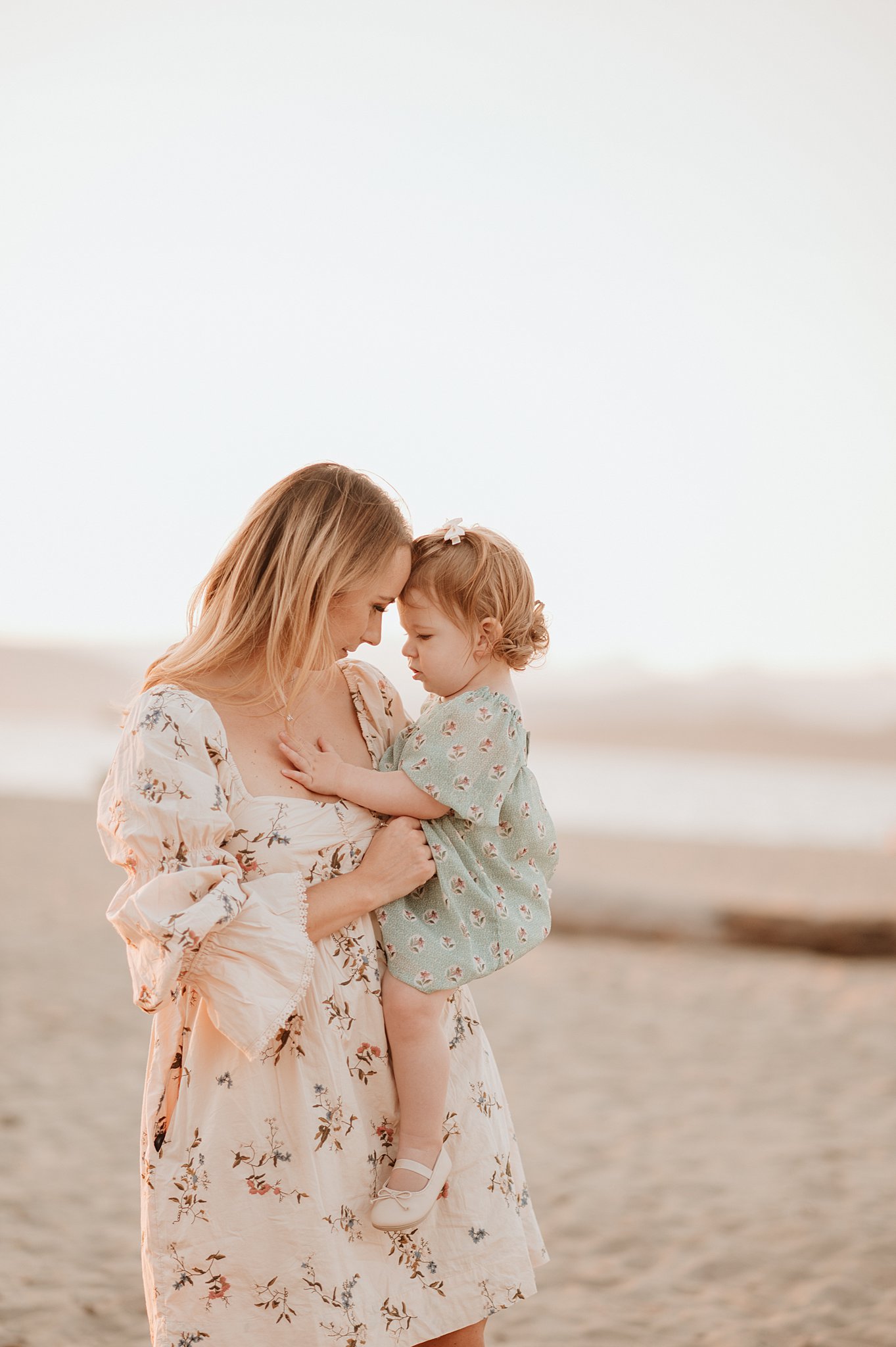 A mother and daughter in floral print dresses stand on a beach at sunset touching foreheads