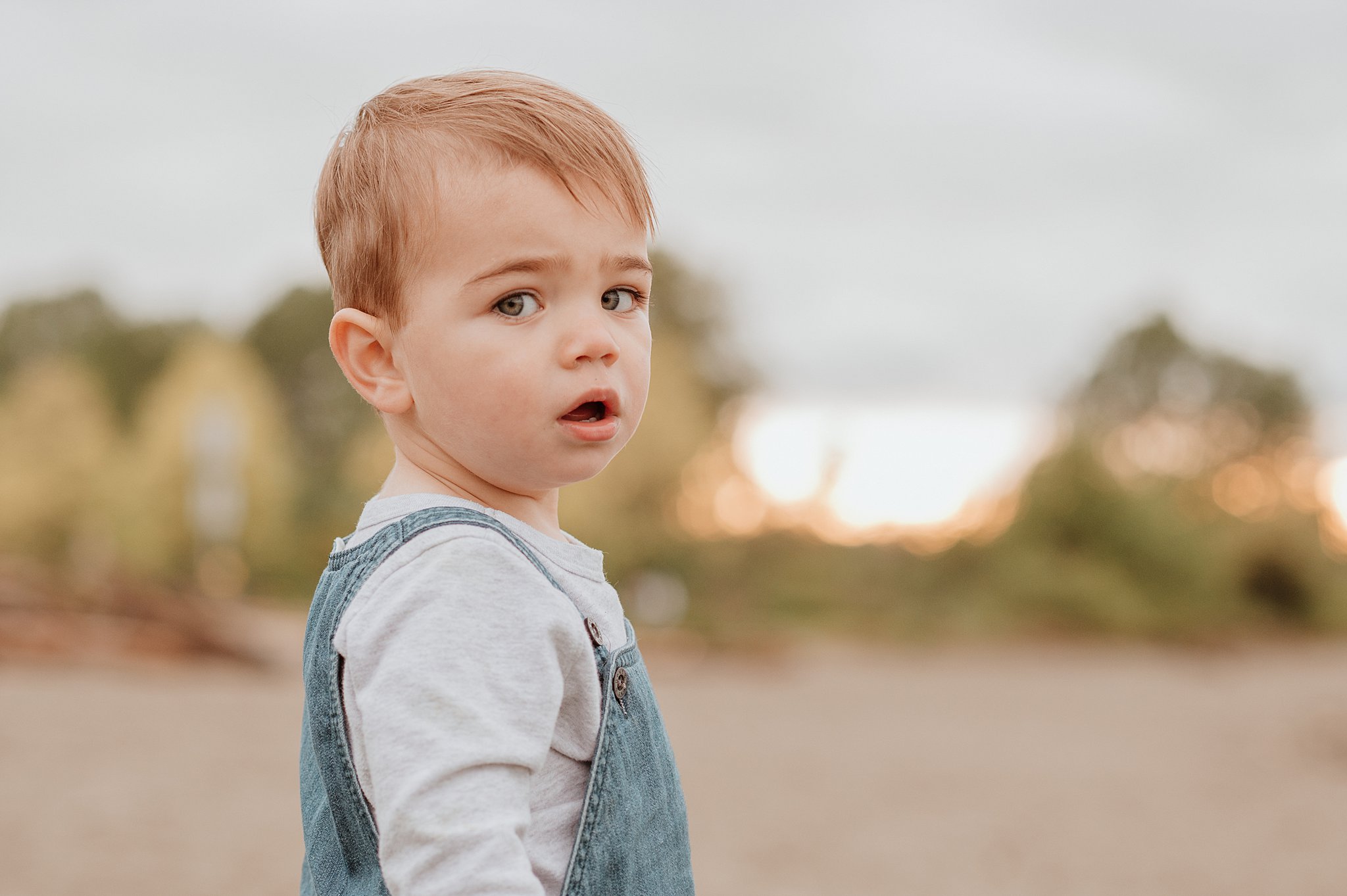 A toddler boy in denim overalls stands on a beach at sunset