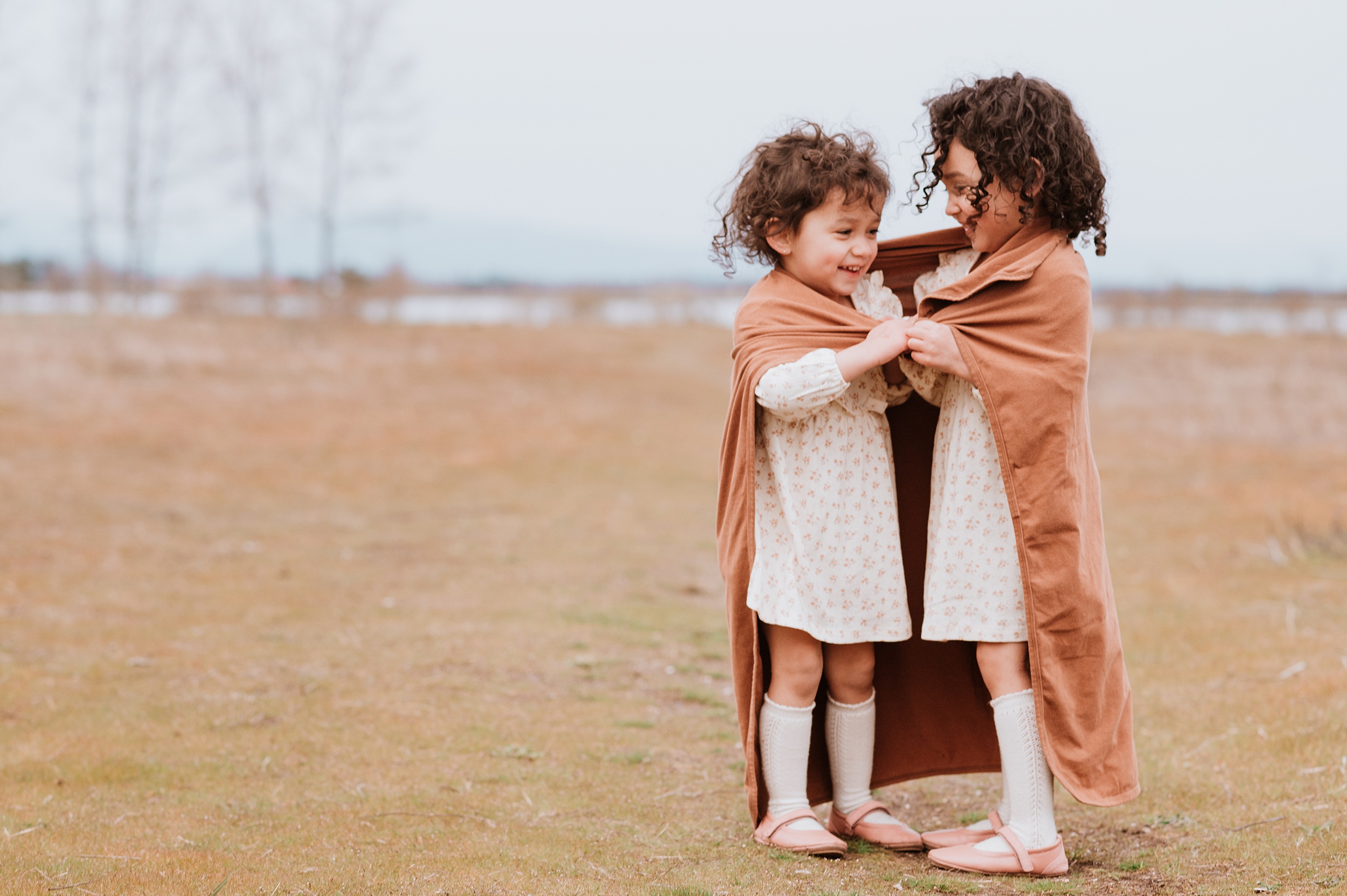 Toddler sisters in matching pink floral dress wrap in a small blanket while laughing in a park after visiting north vancouver pediatric dentist
