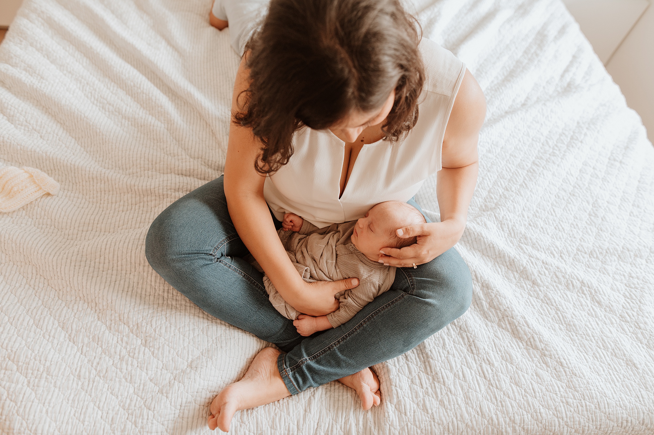 A new mother in jeans and white top cradles her sleeping newborn in her lap while sitting on a bed after visiting north vancouver pediatricians