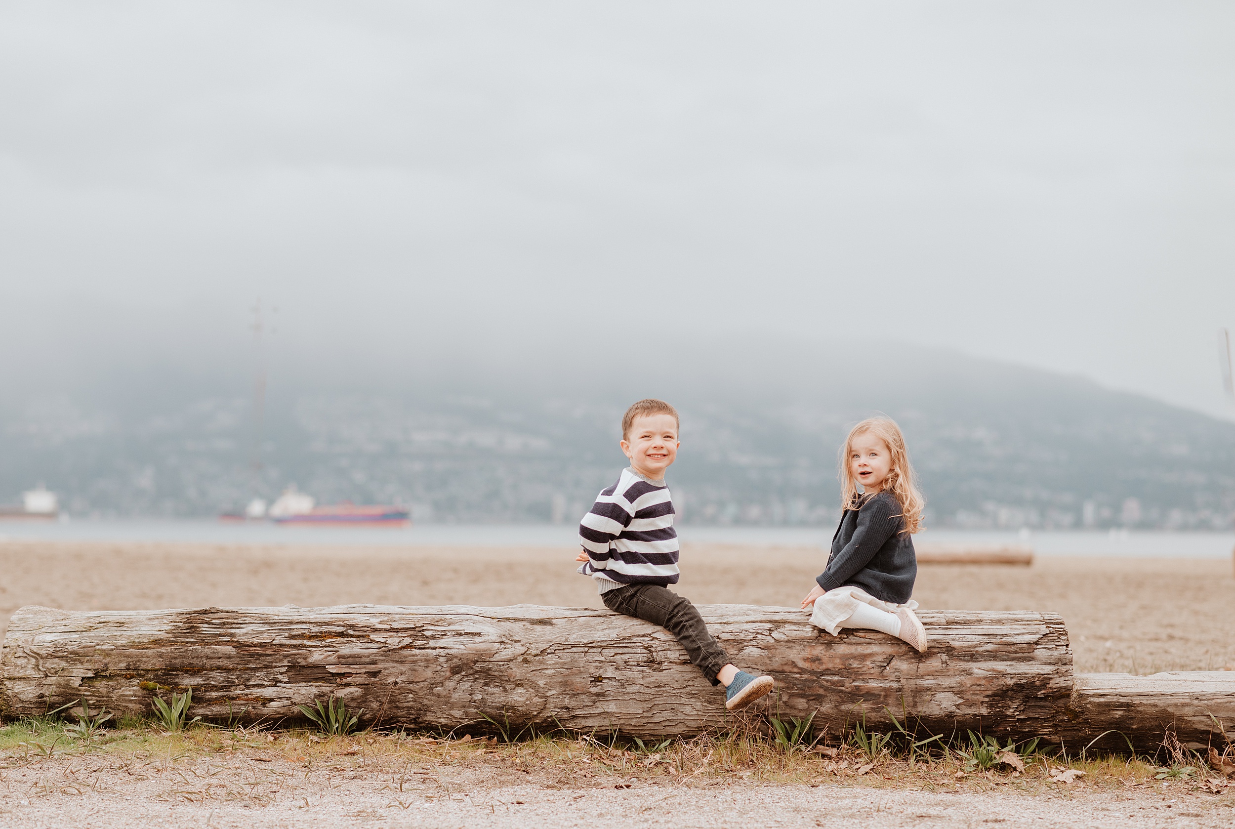 Toddler siblings wearing black and white straddle a log on a beach smiling before meeting vancouver babysitters
