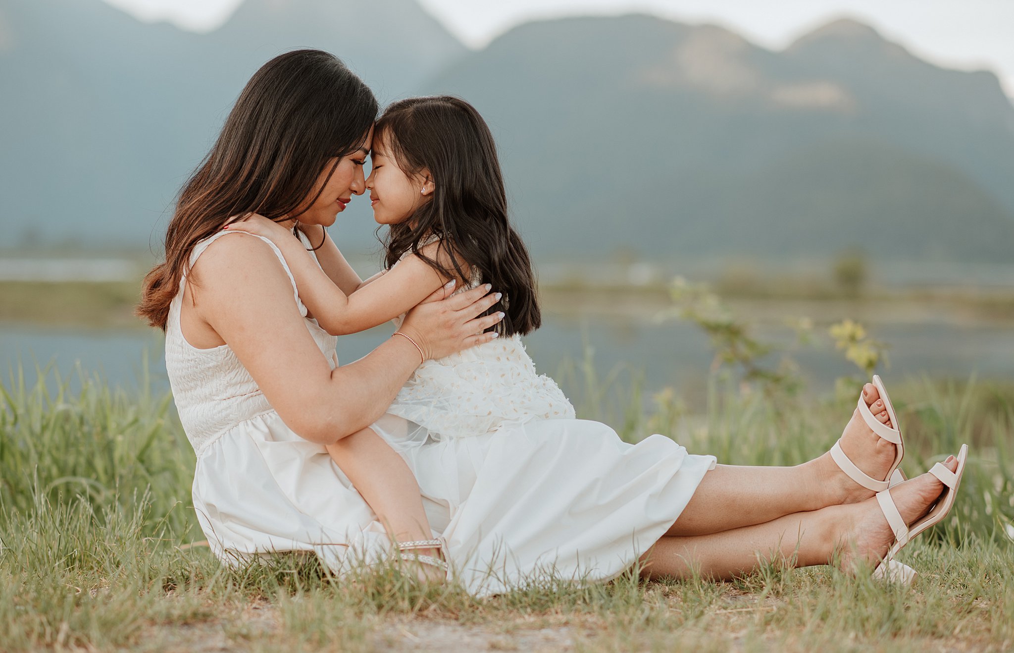 A toddler girl sits in her moms lap by a mountain lake touching noses