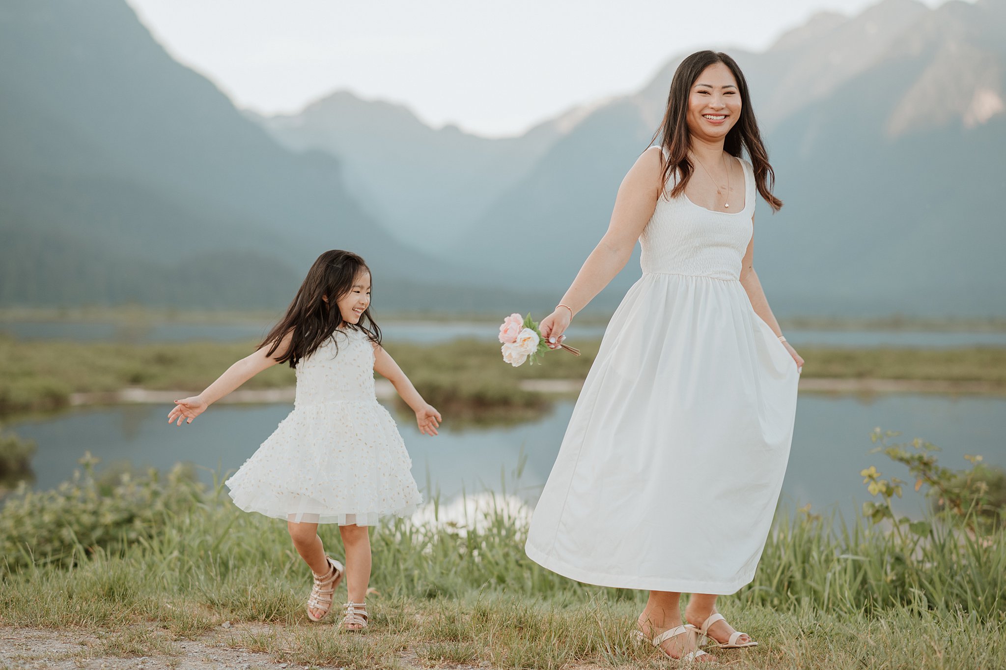A mother and daughter in white dresses play and dance by a mountain lake at sunset before visiting vancouver toy stores