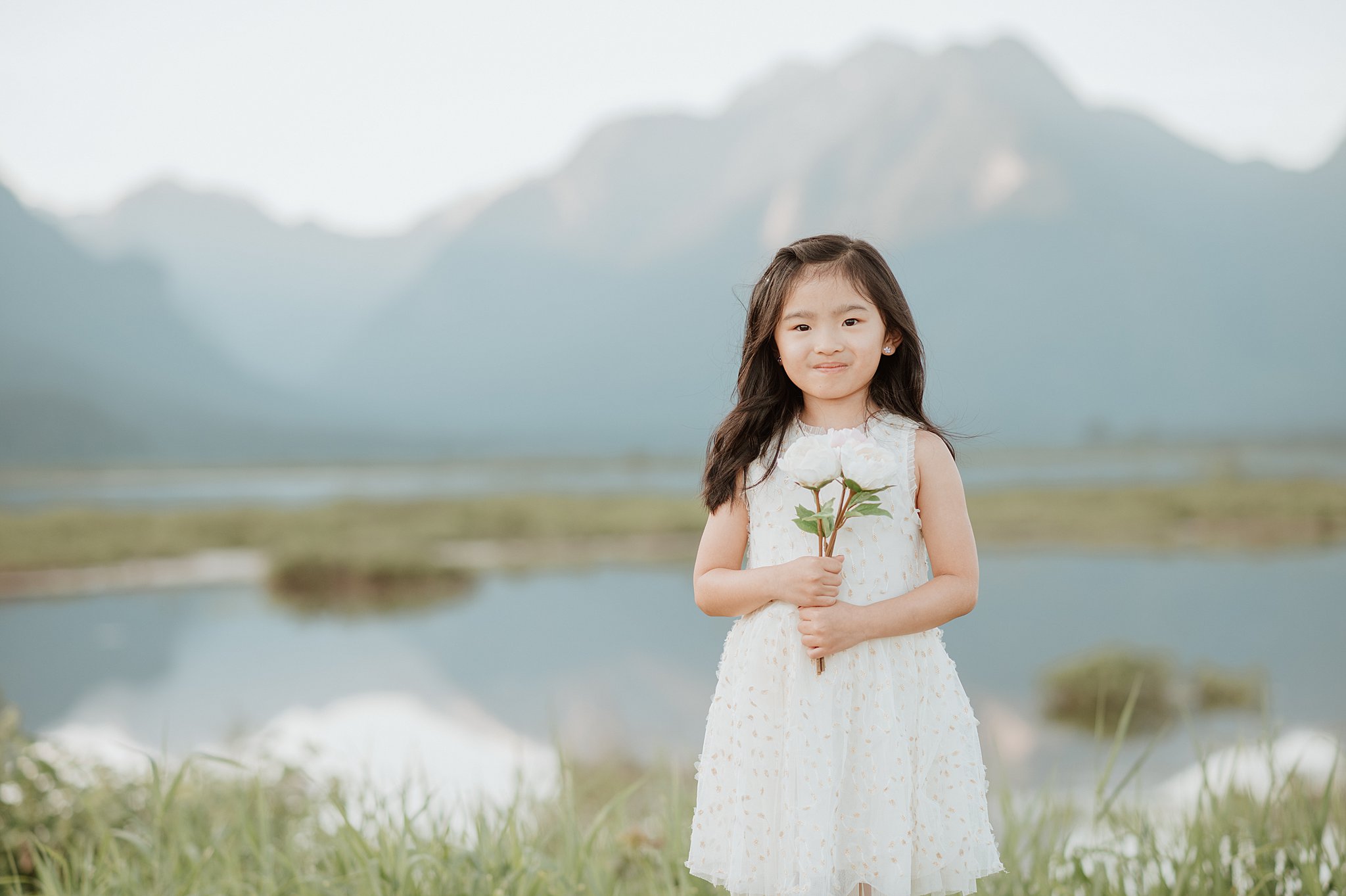 A young girl in a white dress stands by a mountain lake holding two white roses before visiting vancouver toy stores