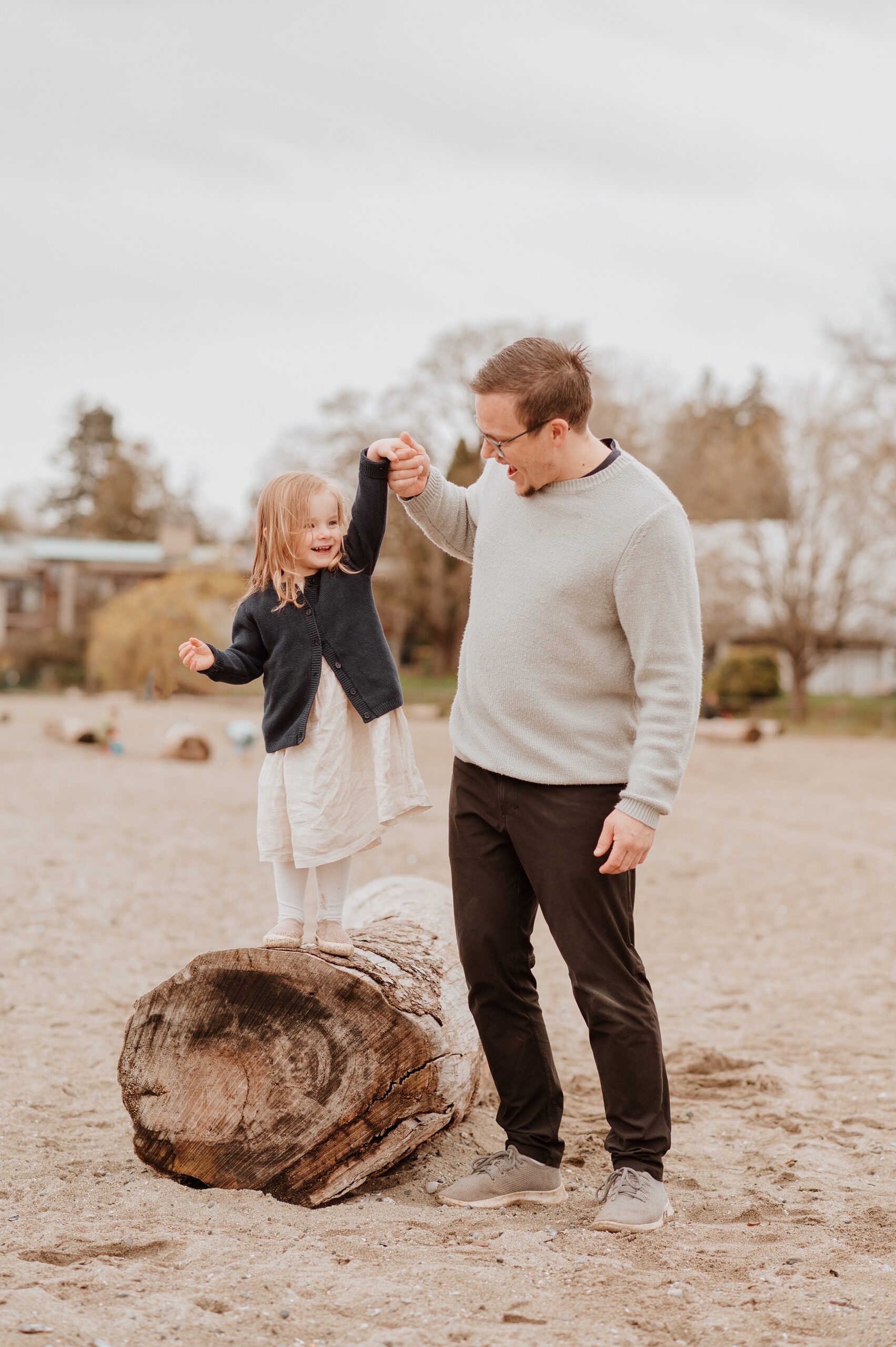 A father plays with his toddler daughter on a piece of beach driftwood in sweaters