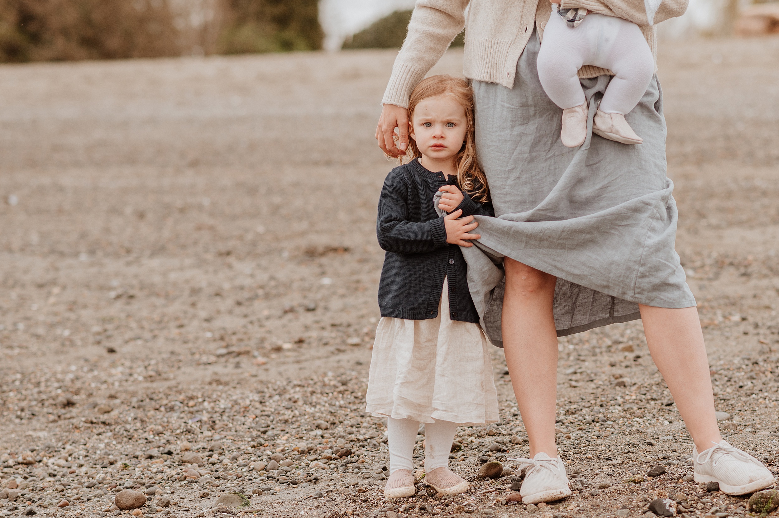 A toddler girl in a dark sweater and white dress holds onto mom's grey dress while standing on a beach before visiting indoor playground vancouver