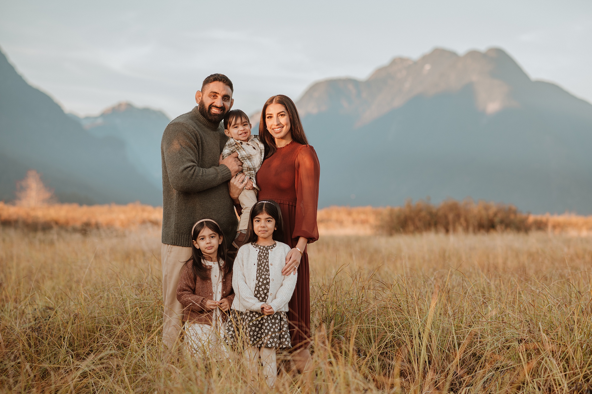 Happy mom and dad stand in a park field surrounded by mountains with their toddler son and two toddler daughters