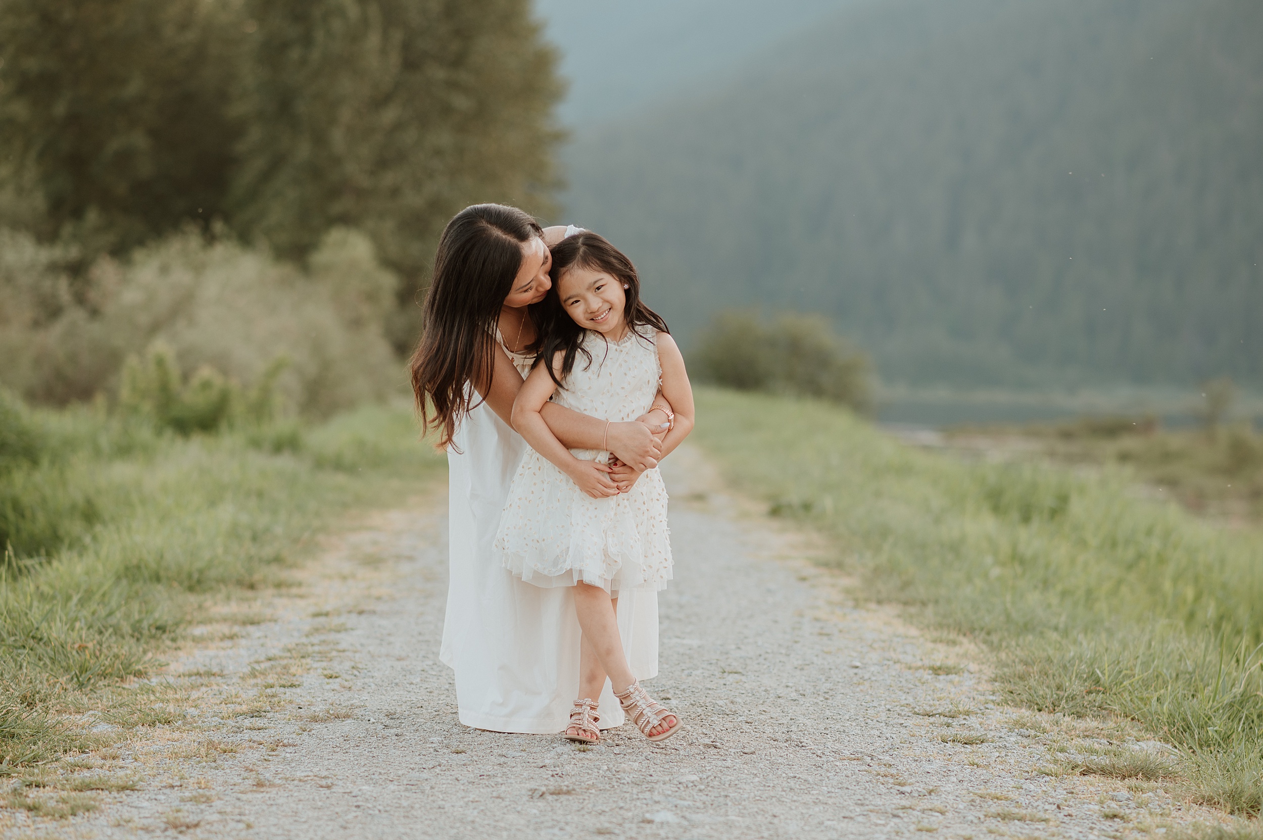 A mother in a white dress hugs onto her toddler daughter as they stand in a park trail by a lake