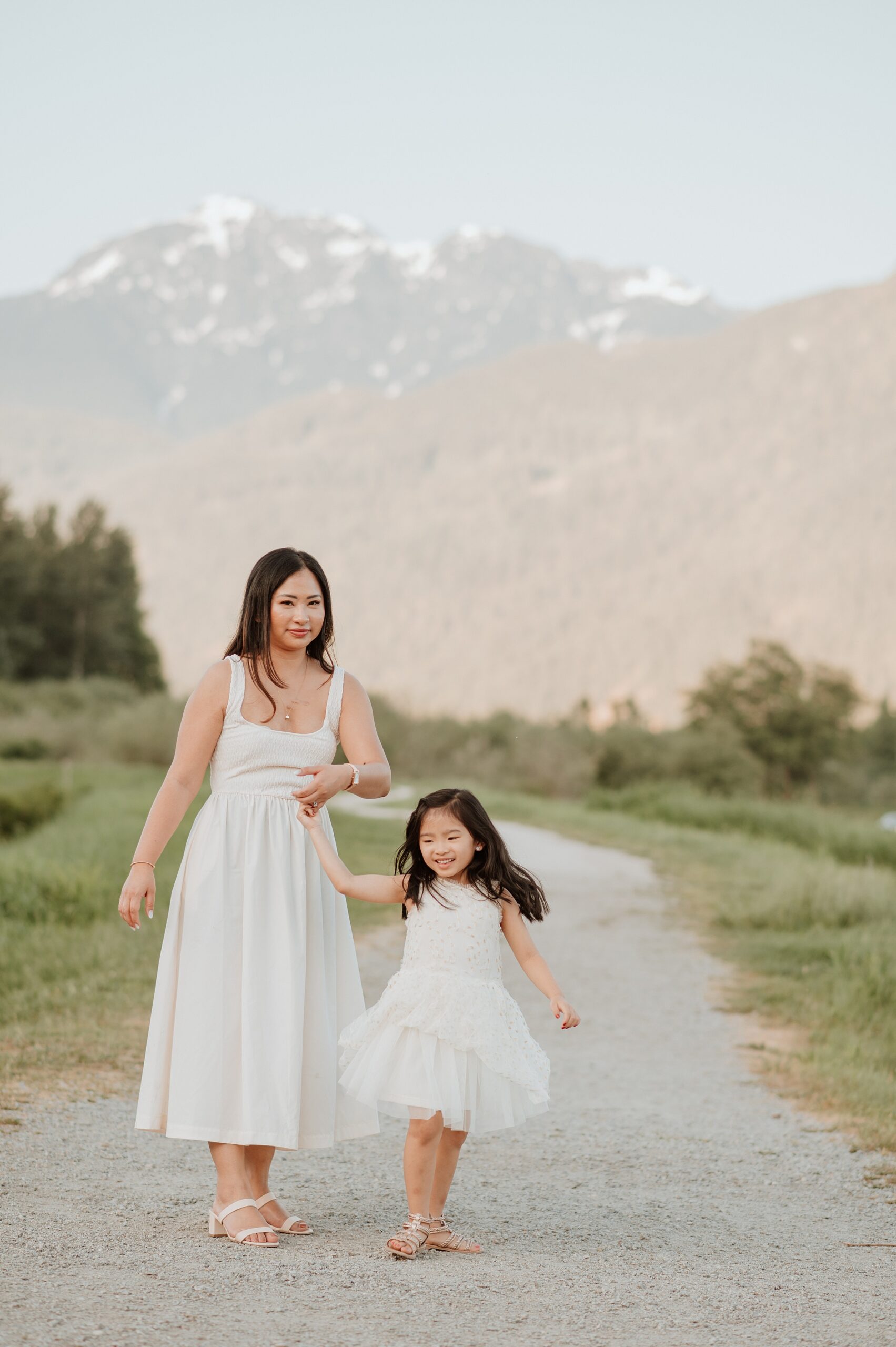A mother in a white dress plays with her toddler daughter in a white dress while walking down a gravel park trail near the mountains before visiting vancouver playgrounds