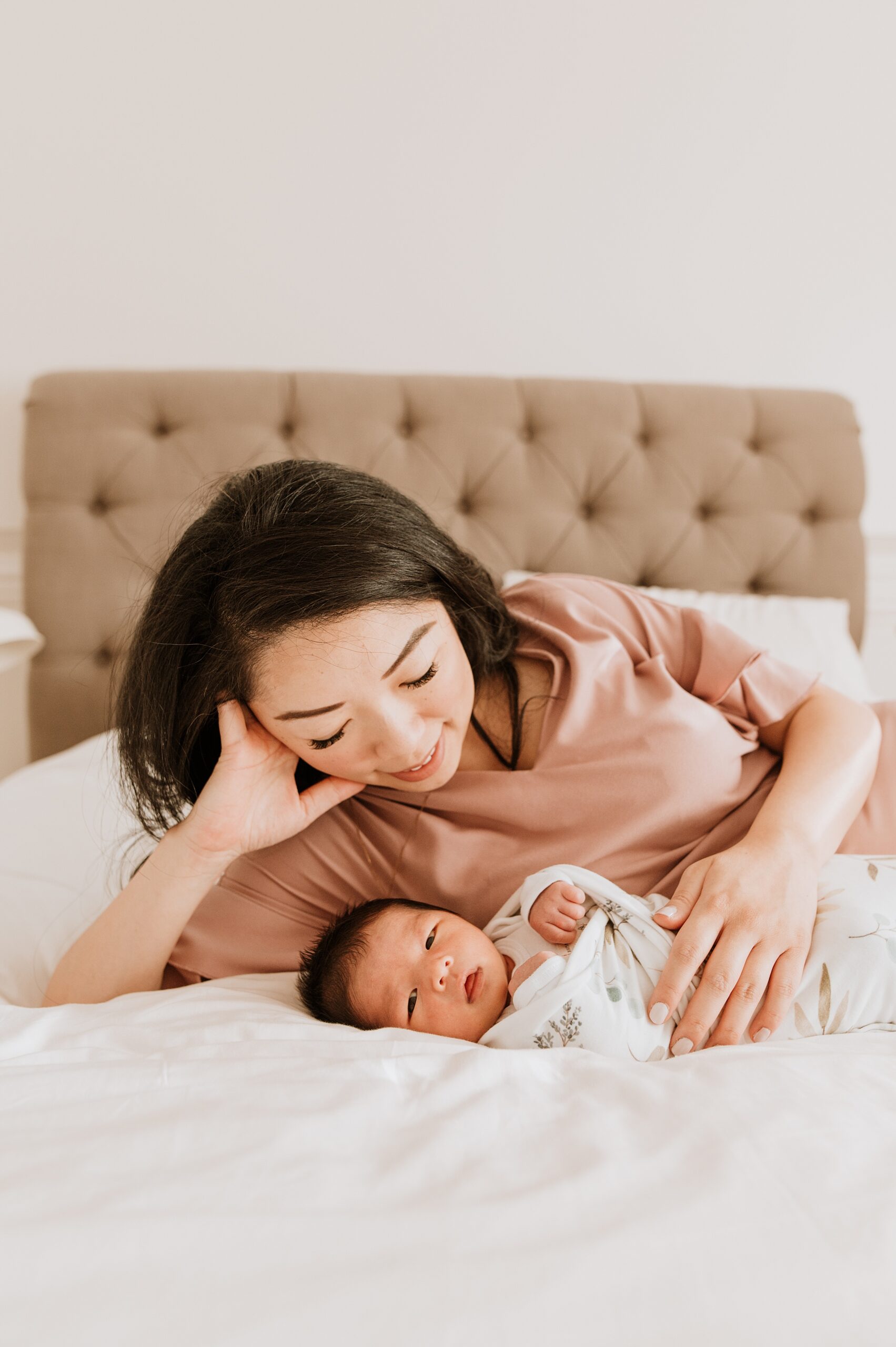 A happy mother snuggles with her newborn baby while laying across a bed