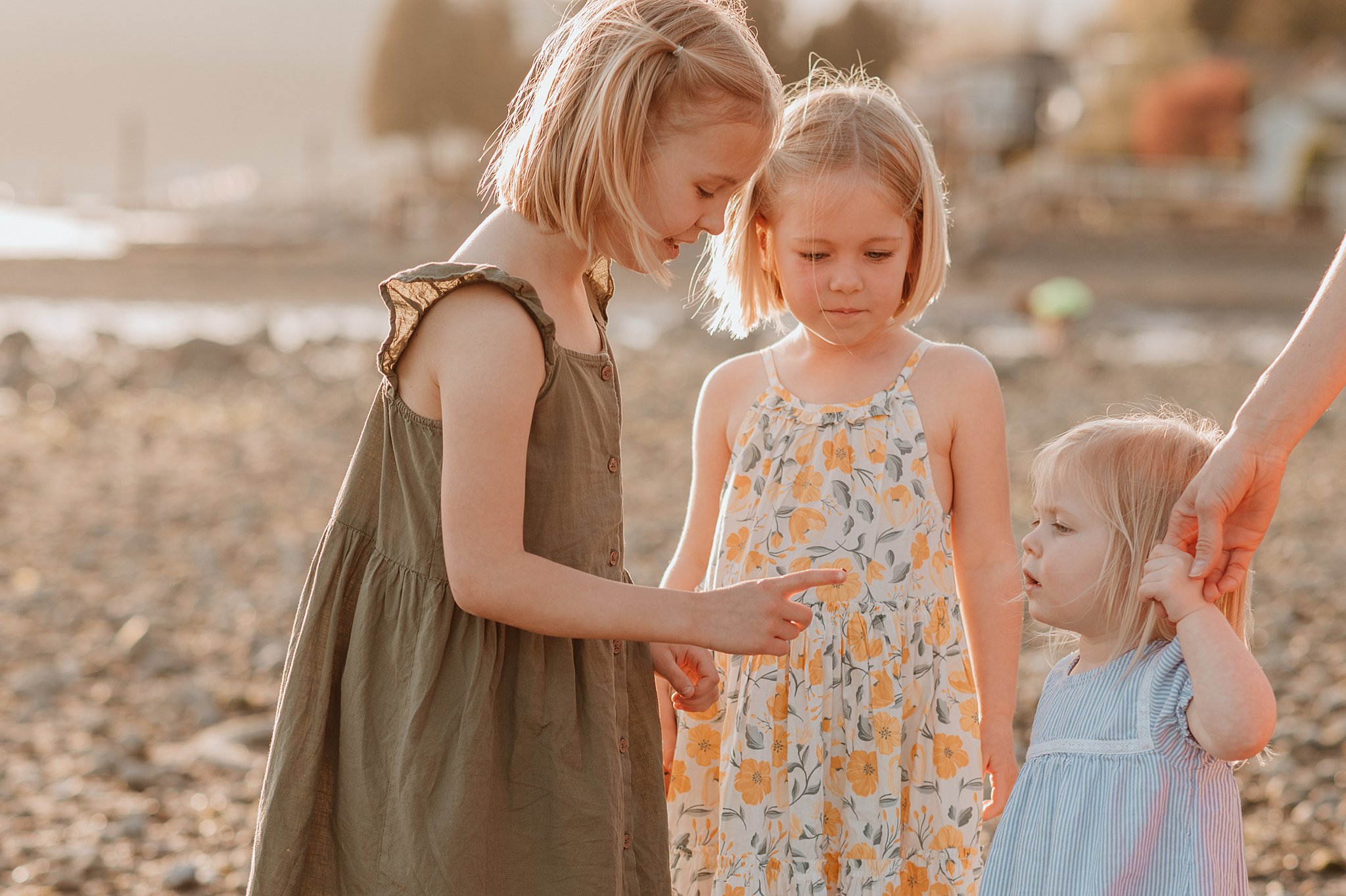 A young girl ina. green dress shows her 2 younger sisters a bug on her finger on a beach at sunset