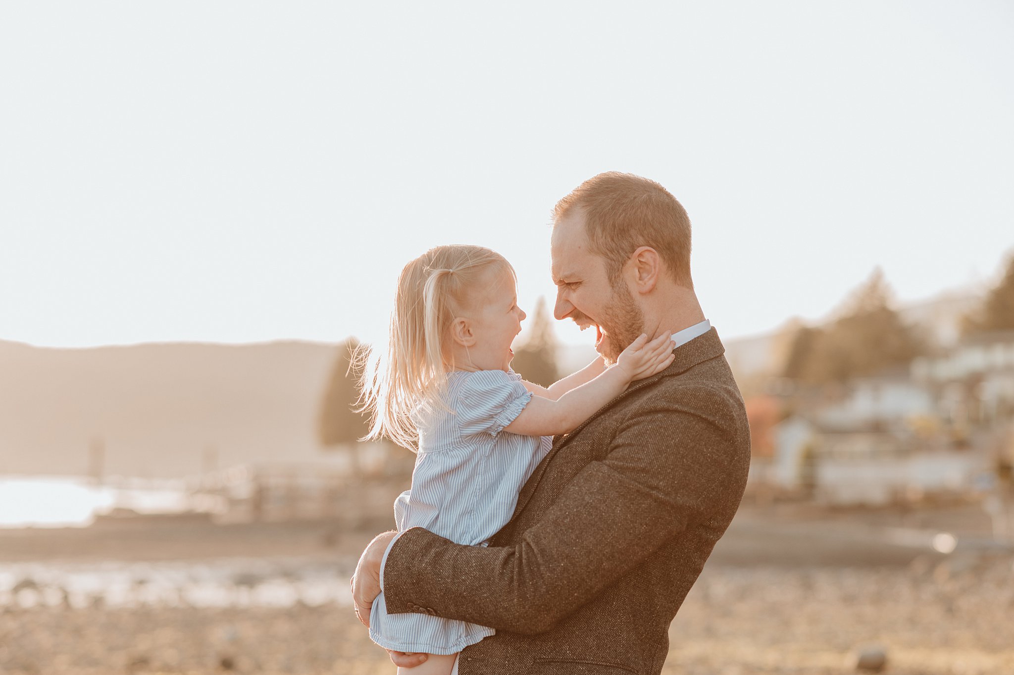 A toddler girl laughs with dad while playing in his arms on a beach at sunset before visiting birthday party venues vancouver