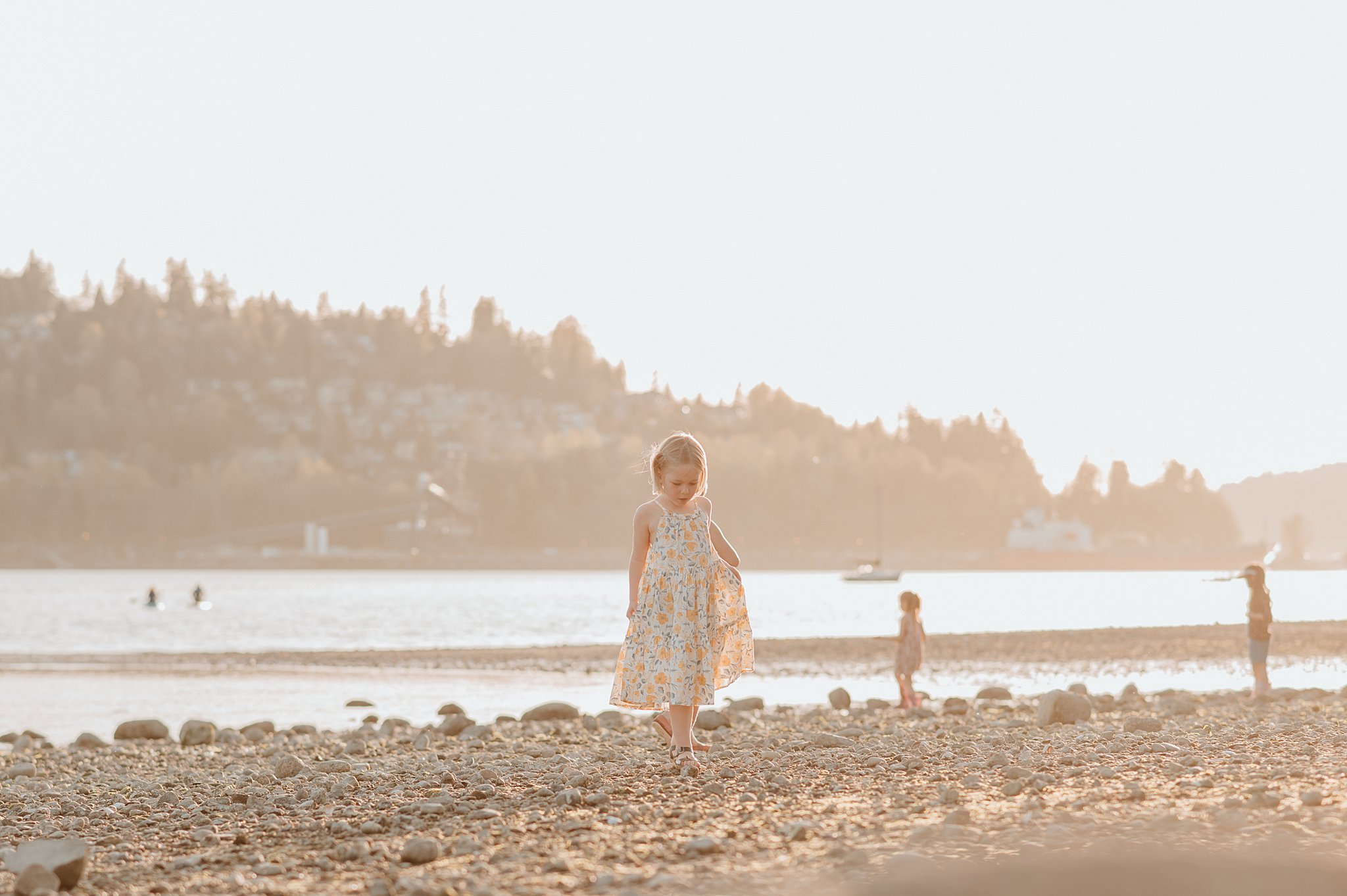 A toddler girl explores a rocky beach at sunset in a yellow sun dress before visiting birthday party venues vancouver