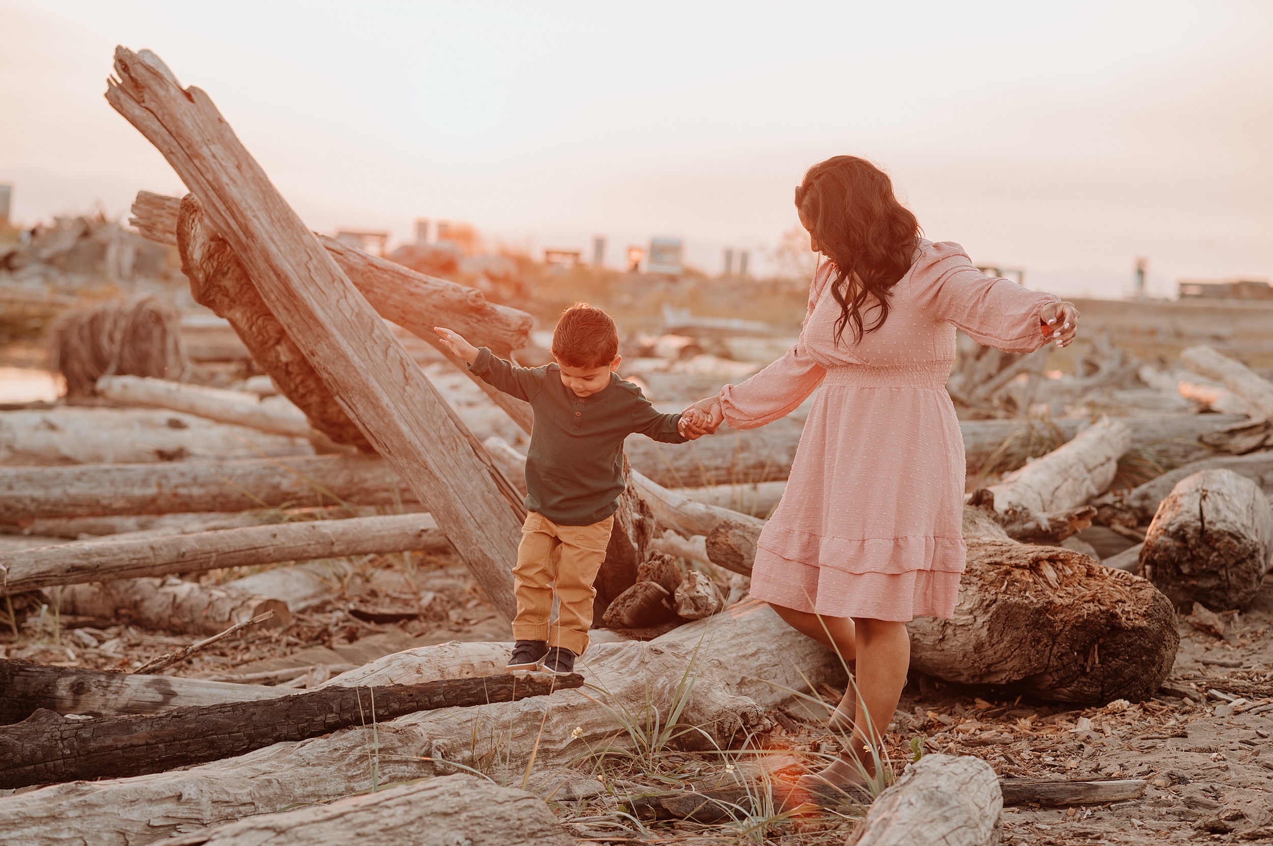 A mother in a pink dress helps her toddler son balance on driftwood logs on a beach on one of the kid friendly hikes vancouver