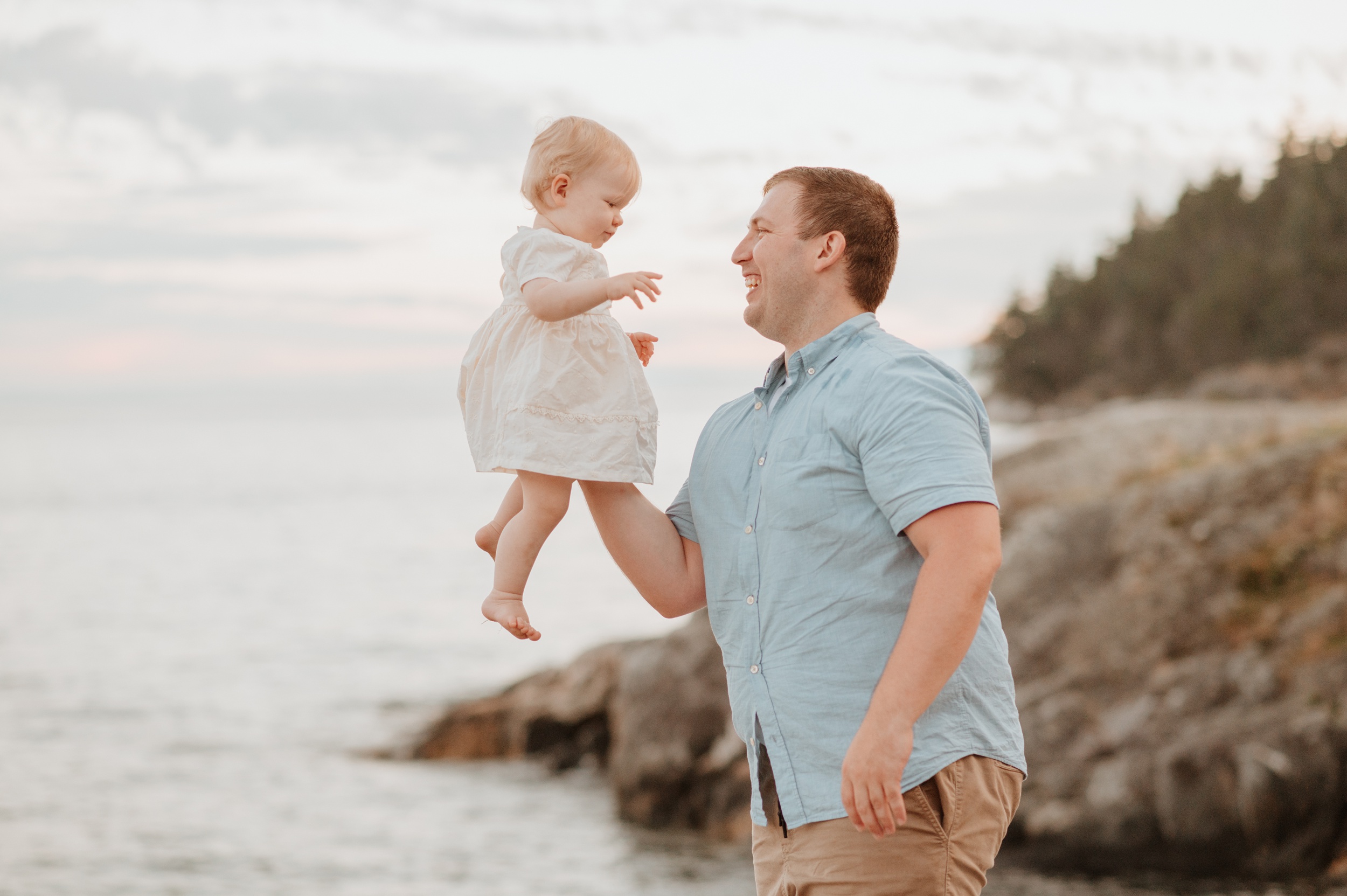 A dad lifts his happy toddler daughter with one arm on a beach at sunset