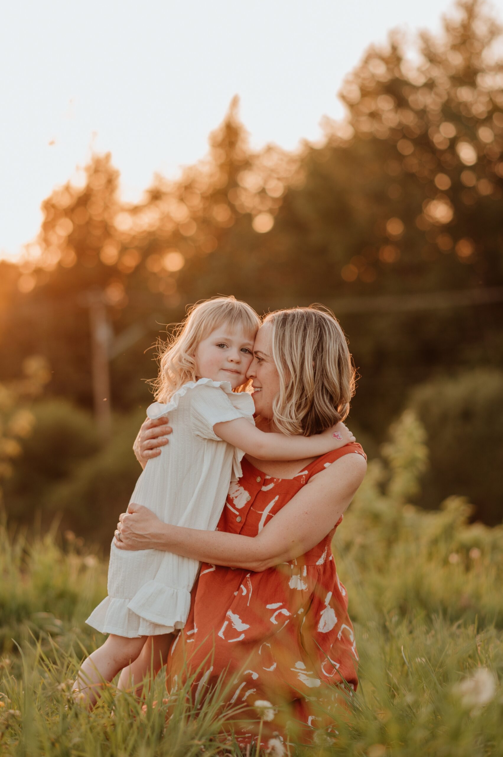 A toddler girl in a white dress hugs mom kneeling in some tall grass at sunset before visiting a vancouver trampoline park