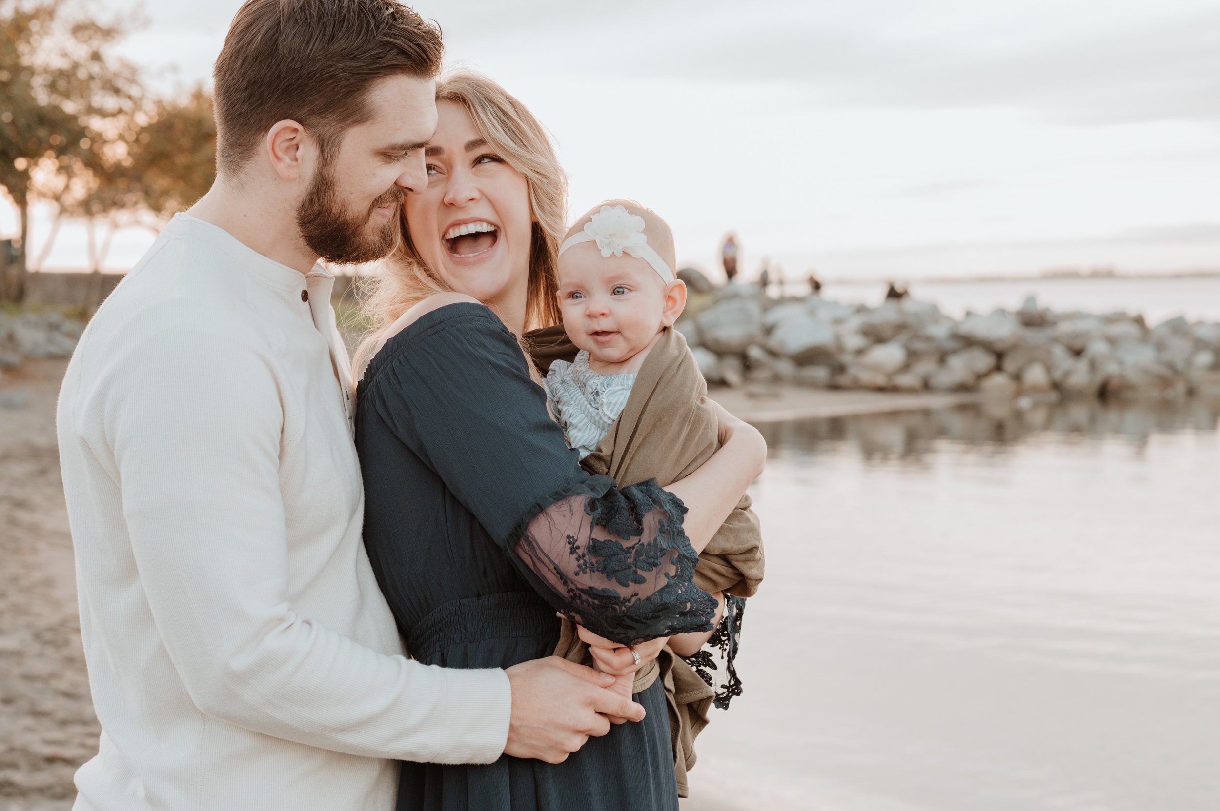 Happy parents stand on a beach by the water laughing with their toddler in mom's arms