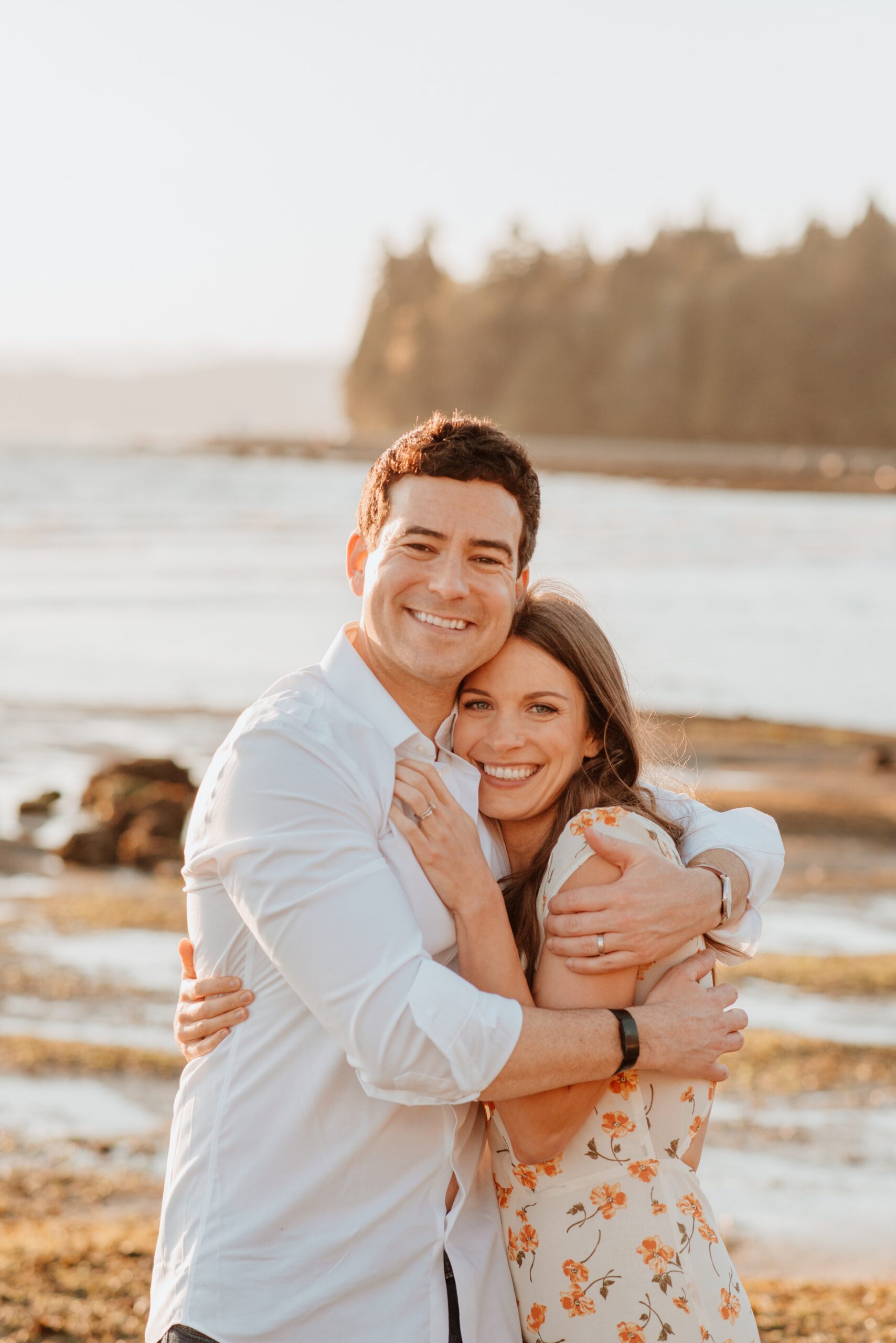 A happy couple hugs while standing on a beach at sunset in a floral print dress and white shirt