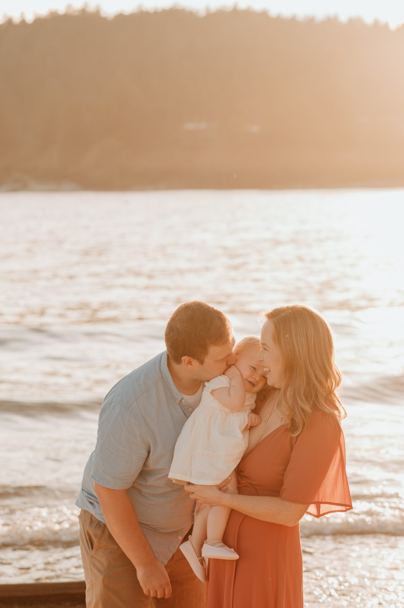 A father kisses his laughing infant in mom's arms while standing on the water at sunset after some parenting classes vancouver