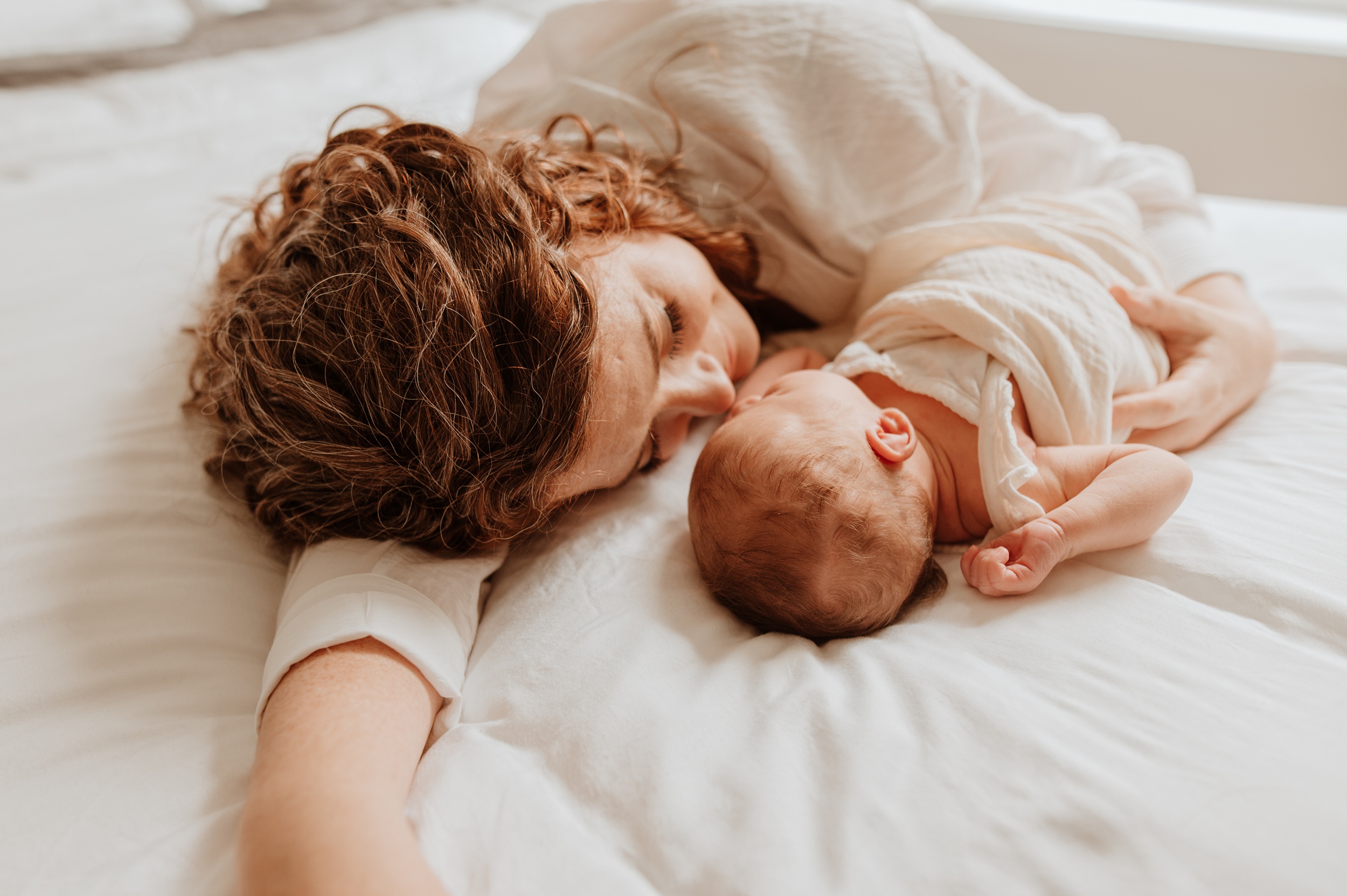 A mother snuggles with her newborn baby on a white bed after meeting vancouver breastfeeding clinic