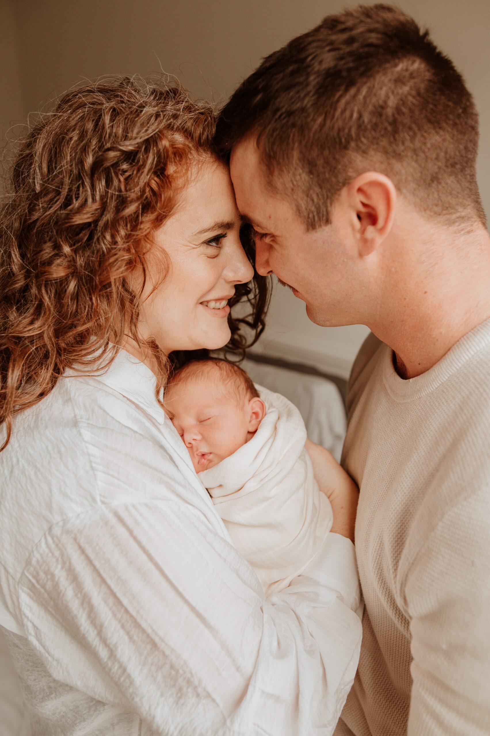 Happy new parents touch foreheads while their newborn sleeps against mom's chest thanks to vancouver breastfeeding clinic