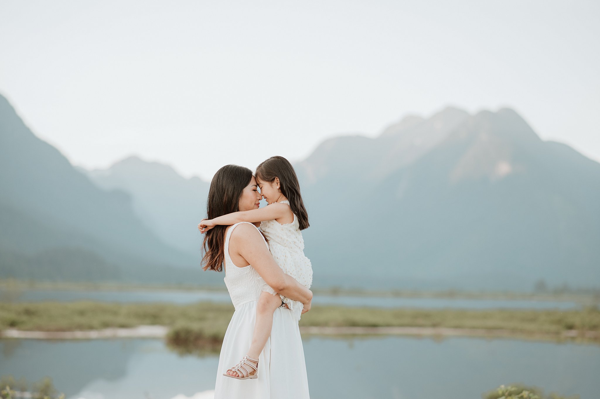 A toddler girl ina. white dress sits in mom's arms by a mountain lake touching foreheads