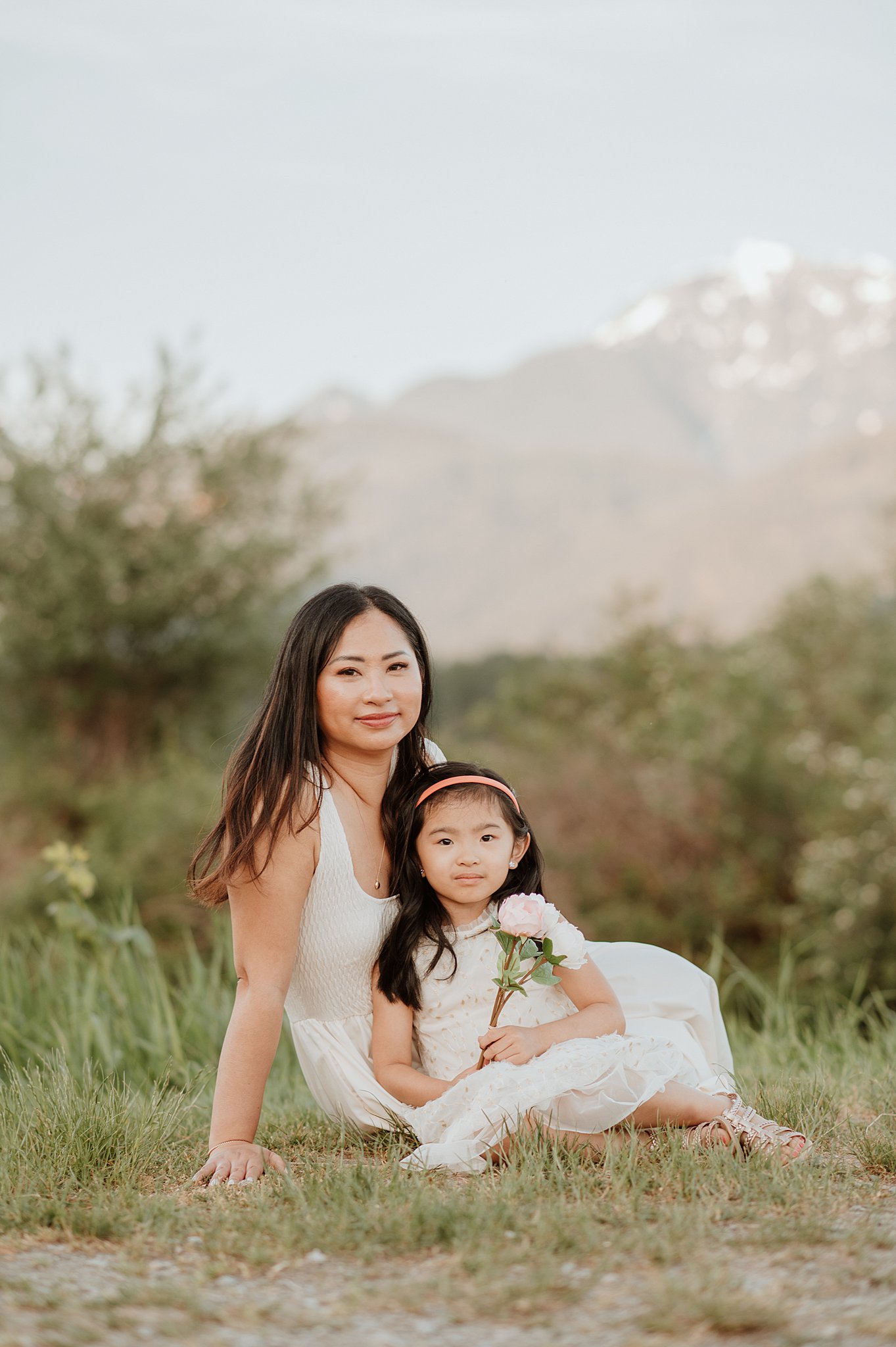 A happy mom in a white dress sits in a park with her toddler daughter in her lap after meeting west coast sitters