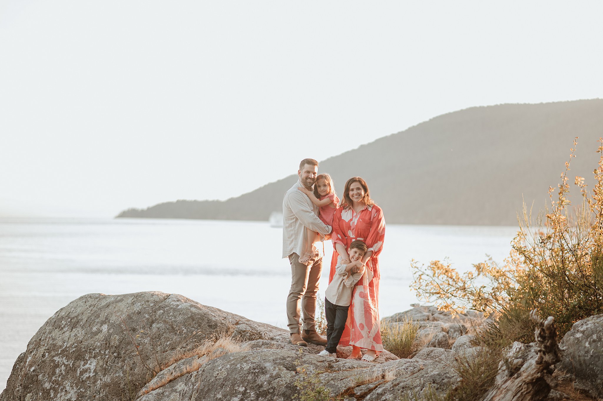 A mom and dad stand on a boulder hugging their toddler son and daughter at sunset