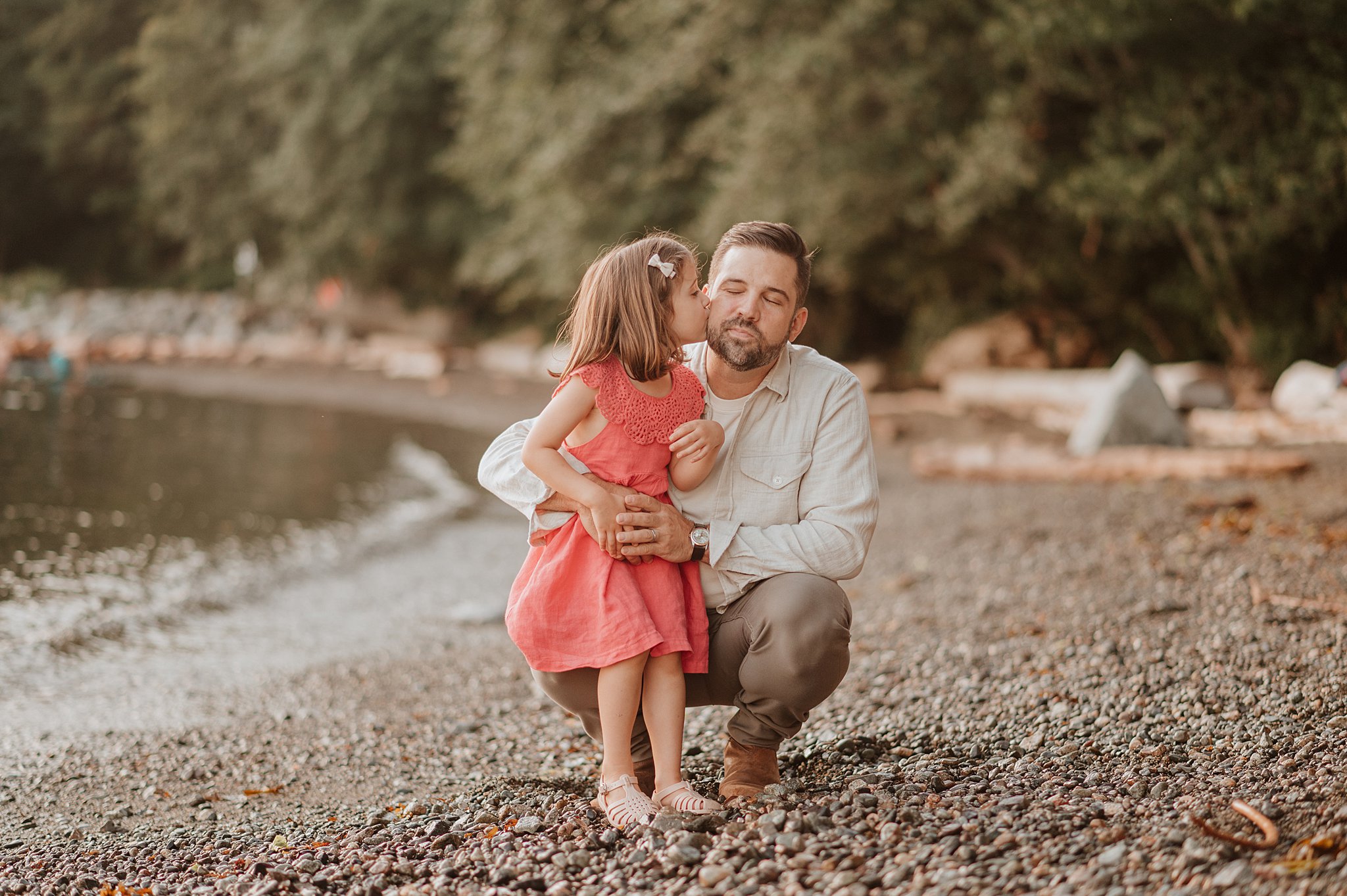 A toddler girl in a pink dress kisses dad's cheek on a beach at sunset after visiting blake and riley