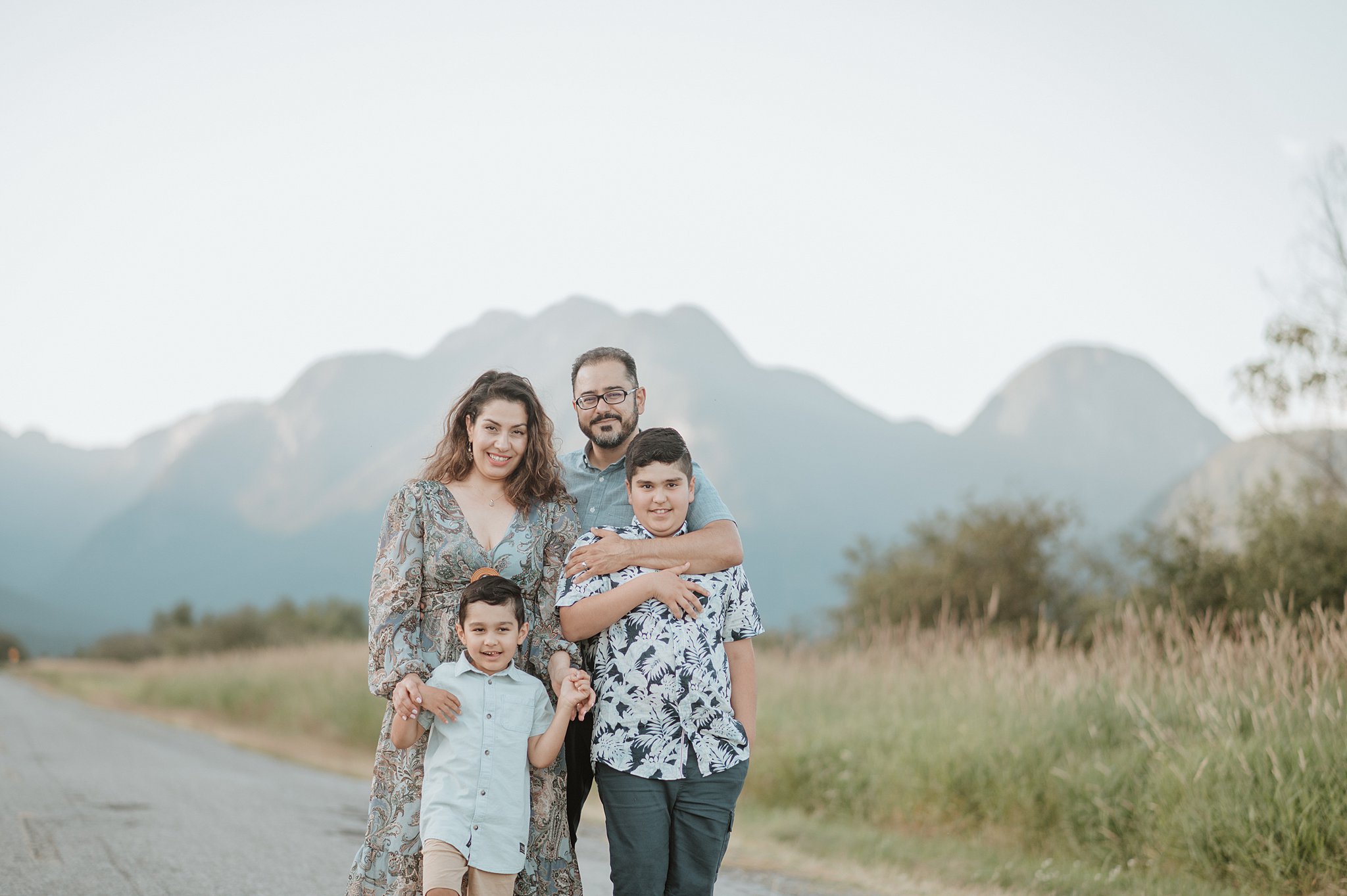 A mom and dad stand hugging their two young sons in a park road at sunset