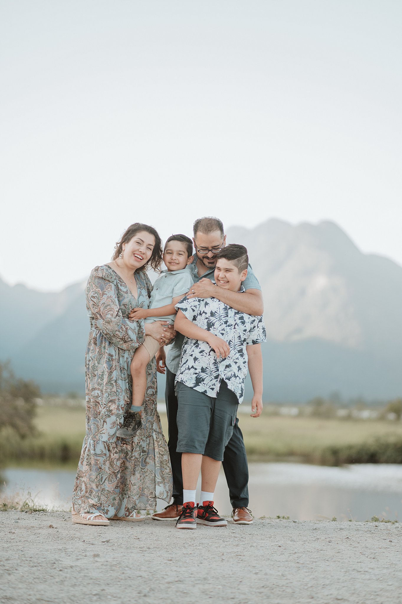 A happy mom and dad hug their toddler and teen sons while standing by a lake at sunset after visiting dilly dally kids