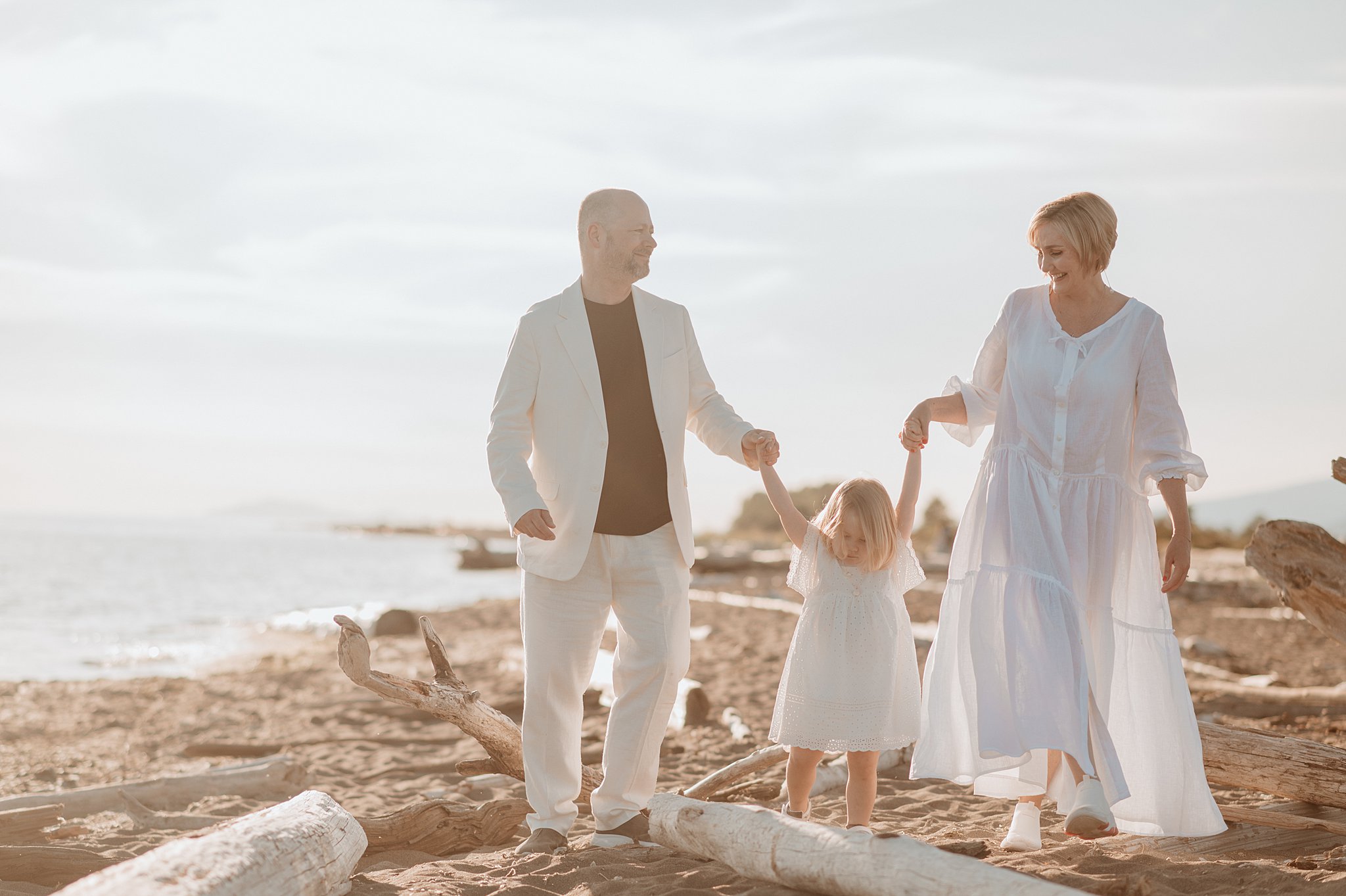 A mom and dad in white walk on a beach helping their toddler daughter in a white dress by holding hands