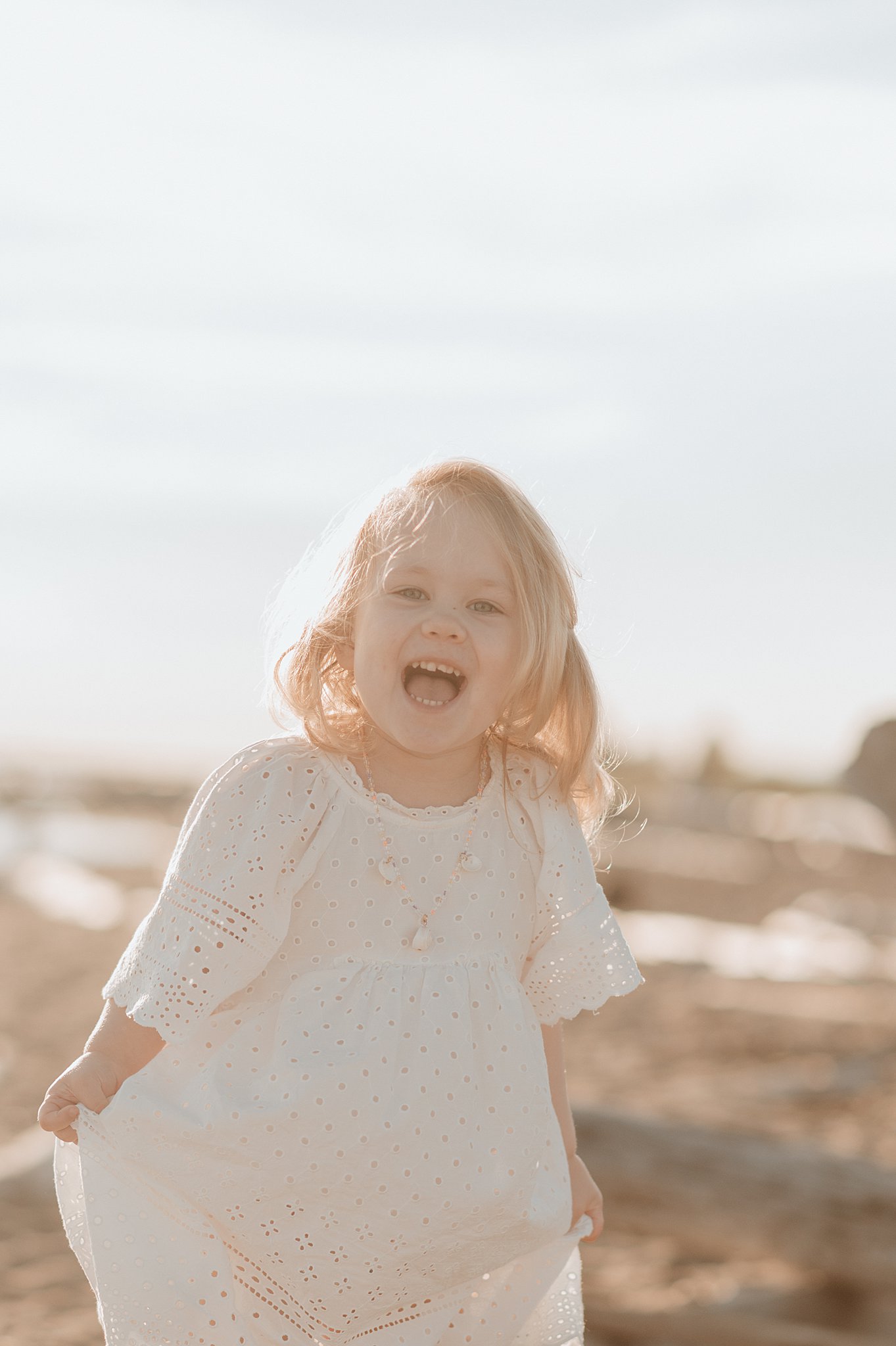 A toddler girl laughs and dances in a white dress on a beach at sunset after visiting isola bella vancouver