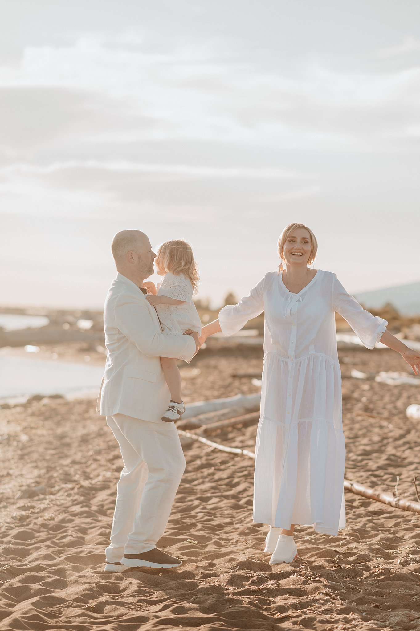 A mom and dad dance on a beach in white at sunset with their toddler daughter in dad's arms after visiting isola bella vancouver