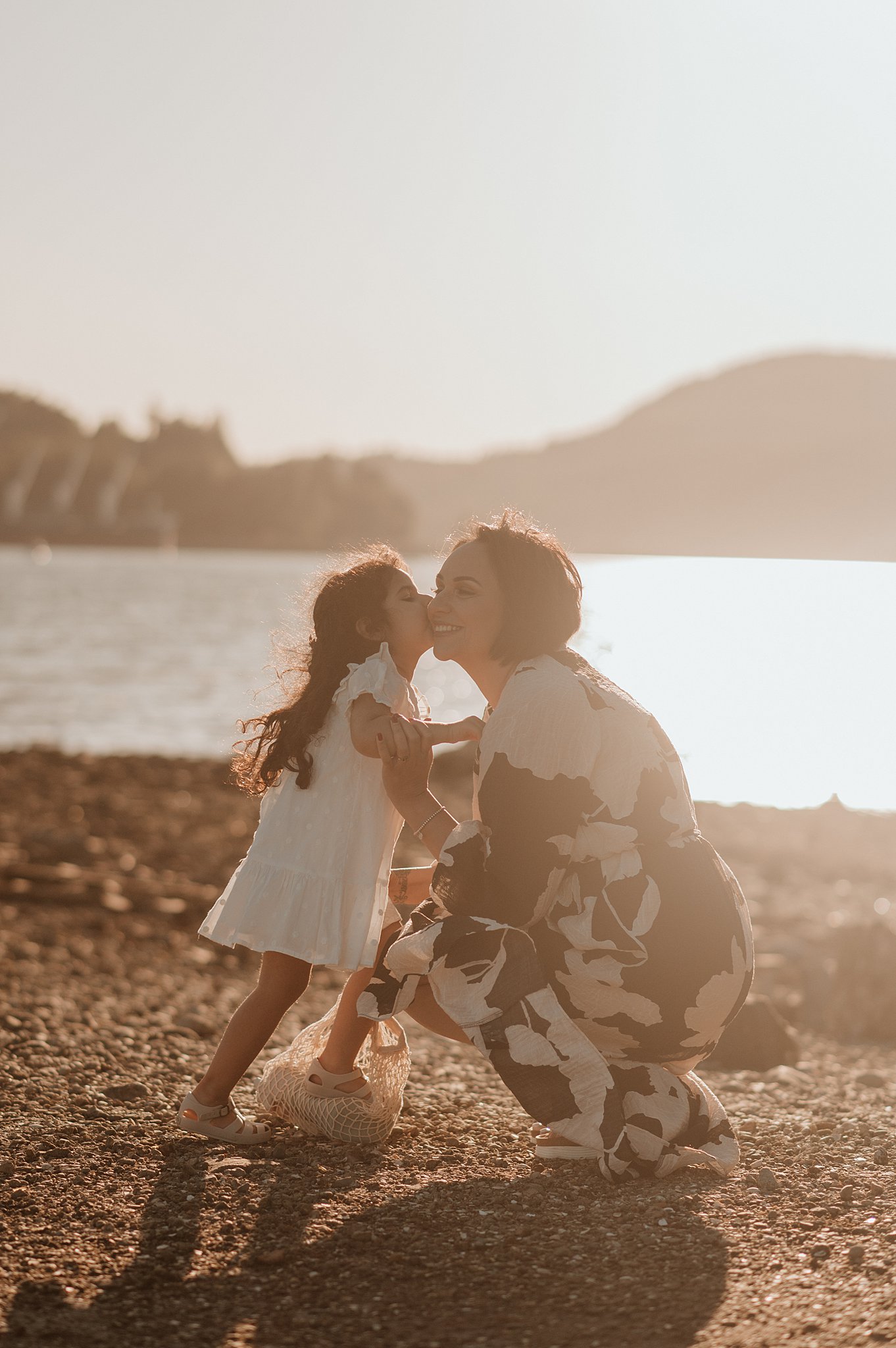 A toddler girl in a white dress kisses her mom on a beach at sunset