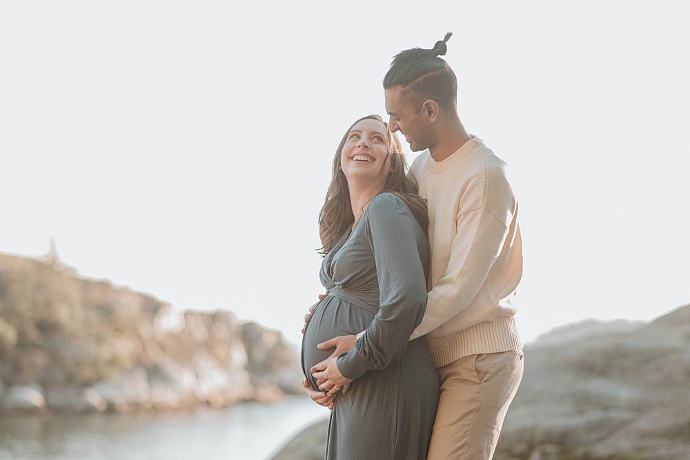 A happy mom to be snuggles against her partner while standing on a rock at susnet