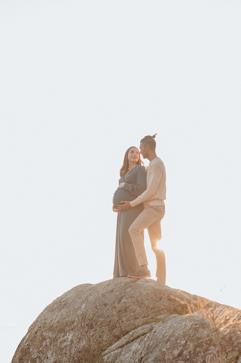 A pregnant woman stands with her partner on a tall rock at sunset while looking at each other after meeting new mummy company