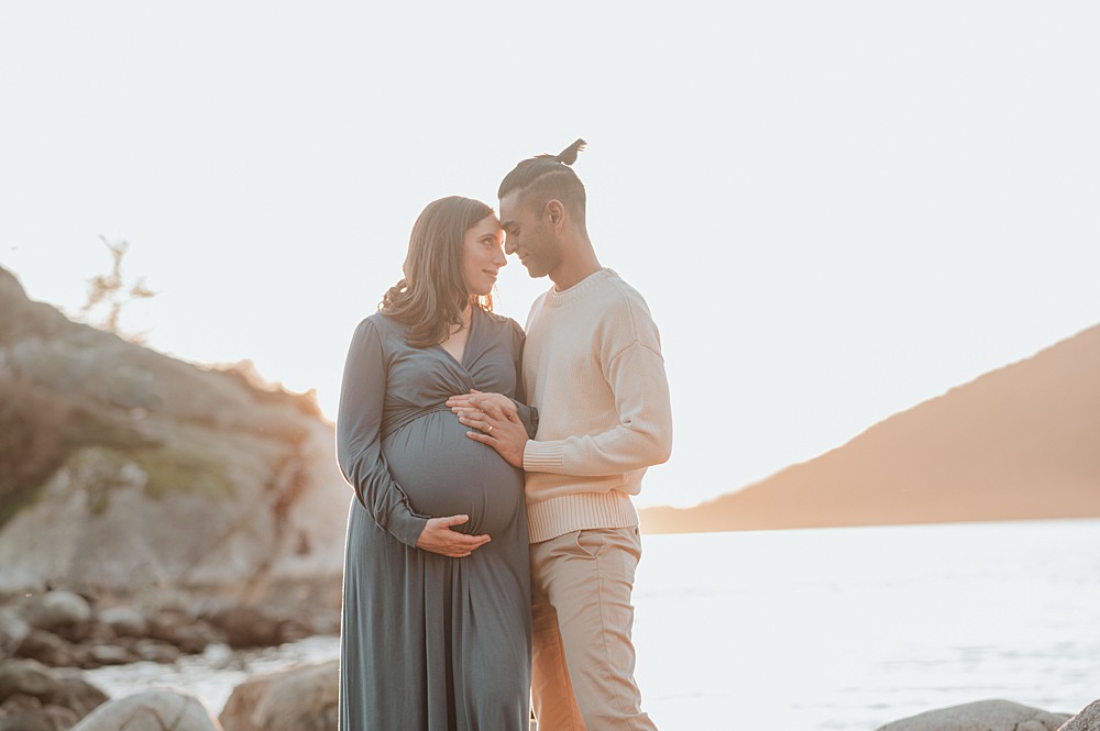 Happy expecting parents snuggle on a rocky beach at sunset while touching the bump after visiting new mummy company