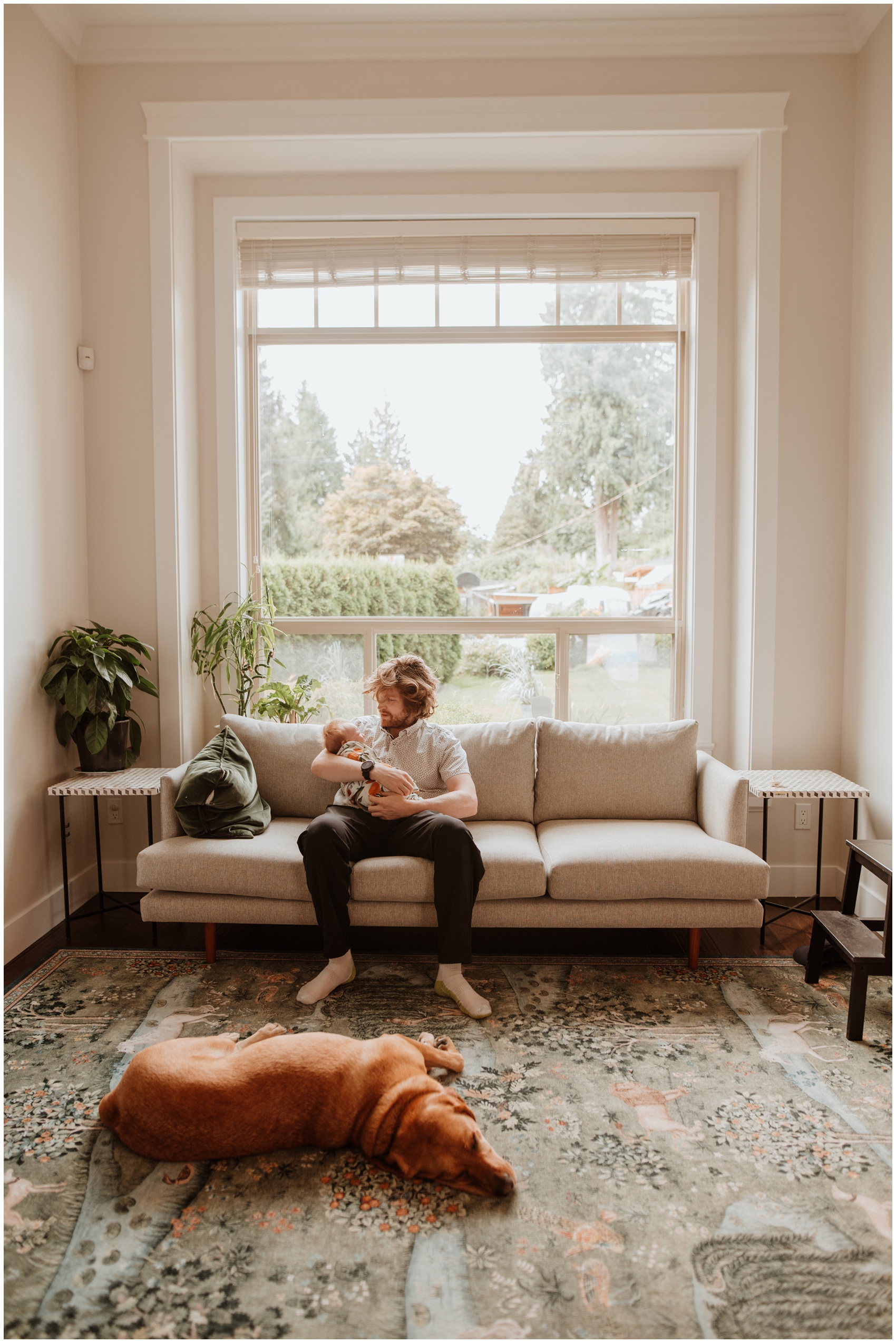 A happy dad sits on a couch under a large window cradling his newborn baby with dog laying at his feet