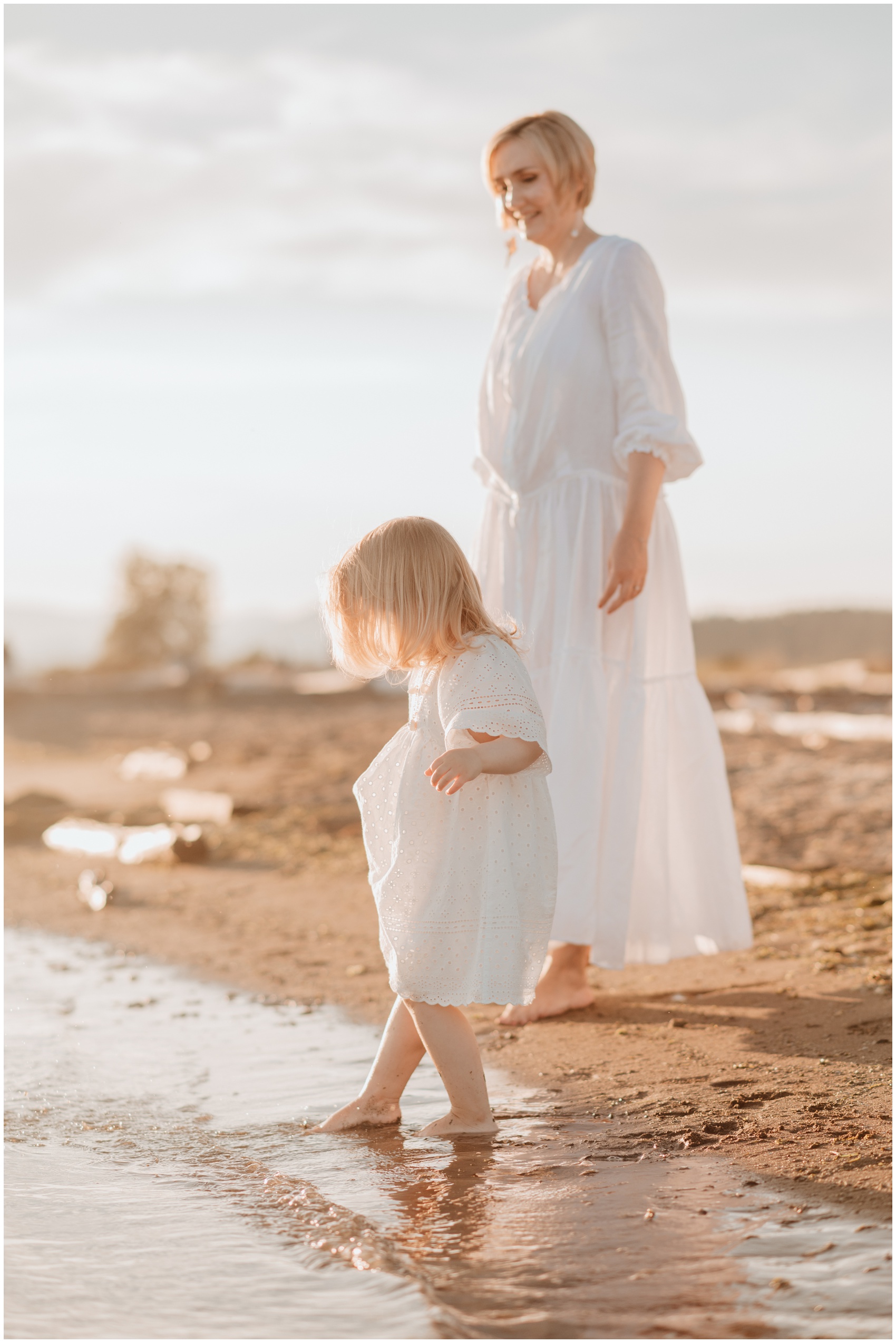 A toddler girl and mom in white dresses play in the water on the beach at sunset