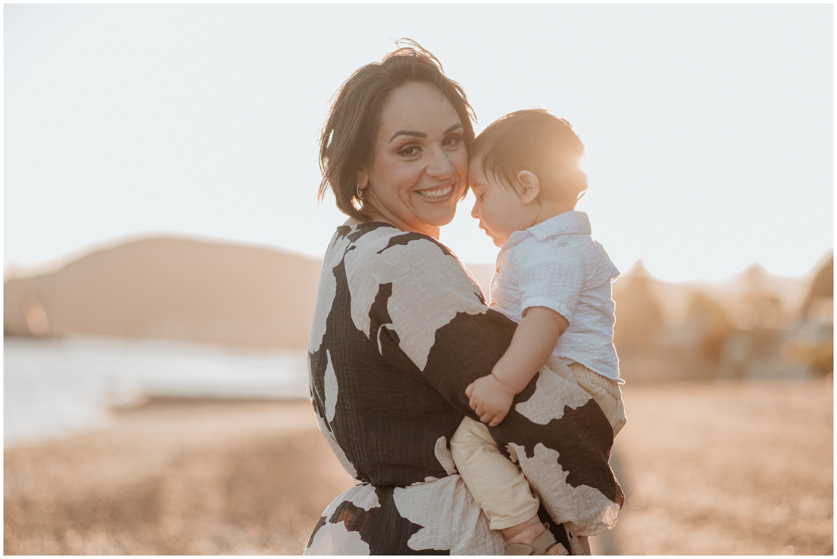 A happy mom smiles over her shoulder while exploring a beach with her toddler in her arms at sunset after meeting the nanny solution