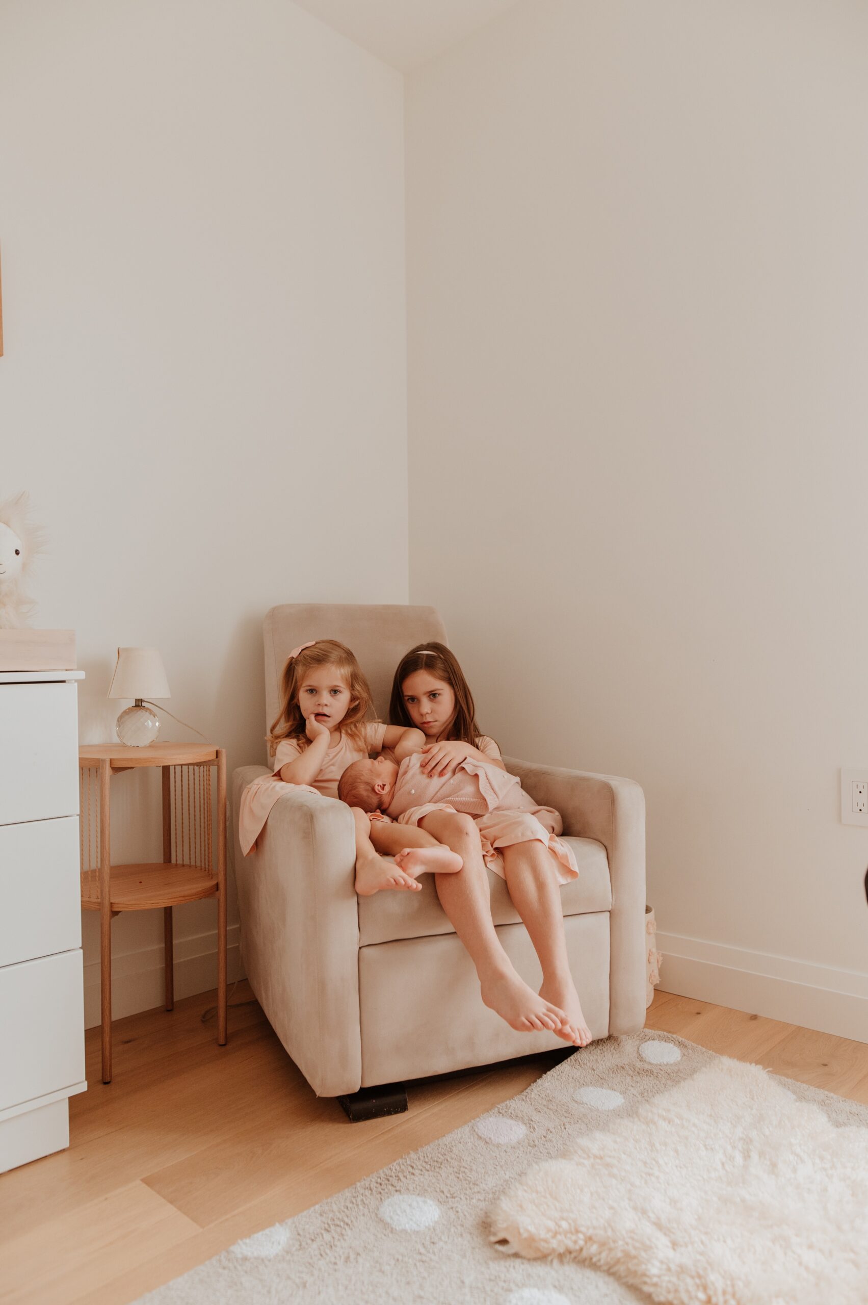 Two young sisters sit in a nursing chair with their newborn baby sister in their lap