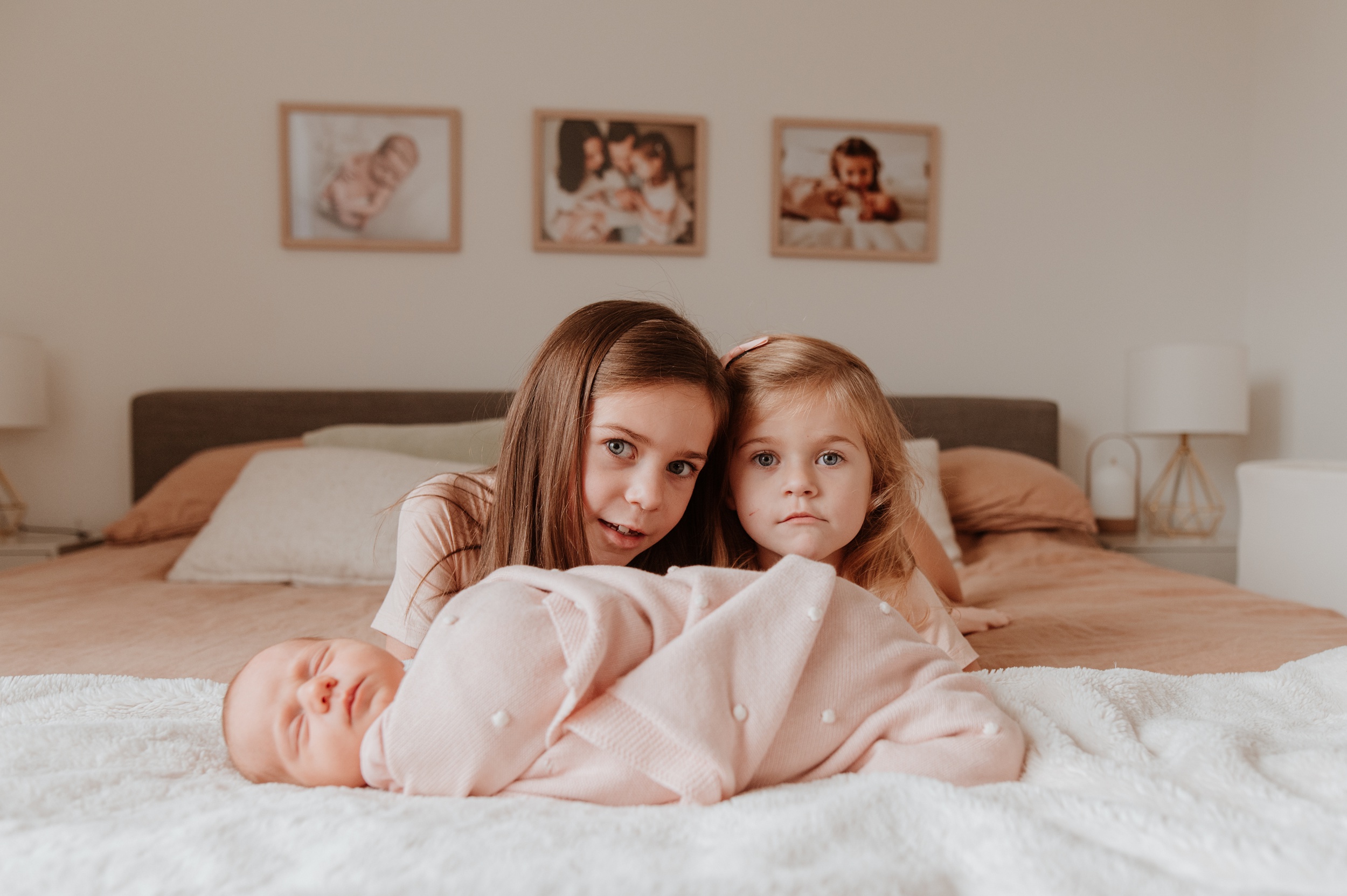 Two toddler sisters lay on a bed behind their newborn sibling after attending tots and tunes