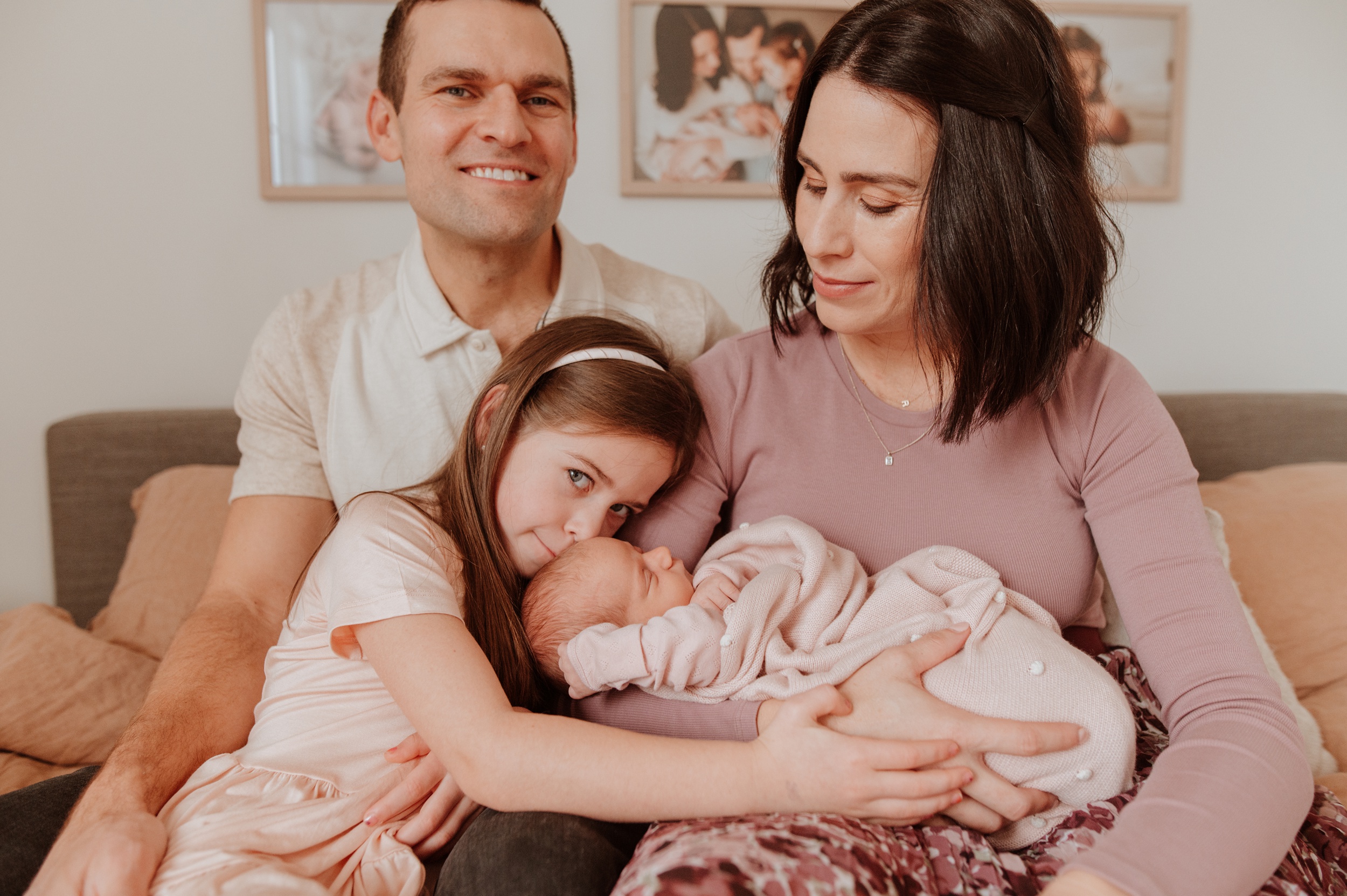 A toddler girl snuggles on her newborn sibling in mom's lap with dad smiling behind them